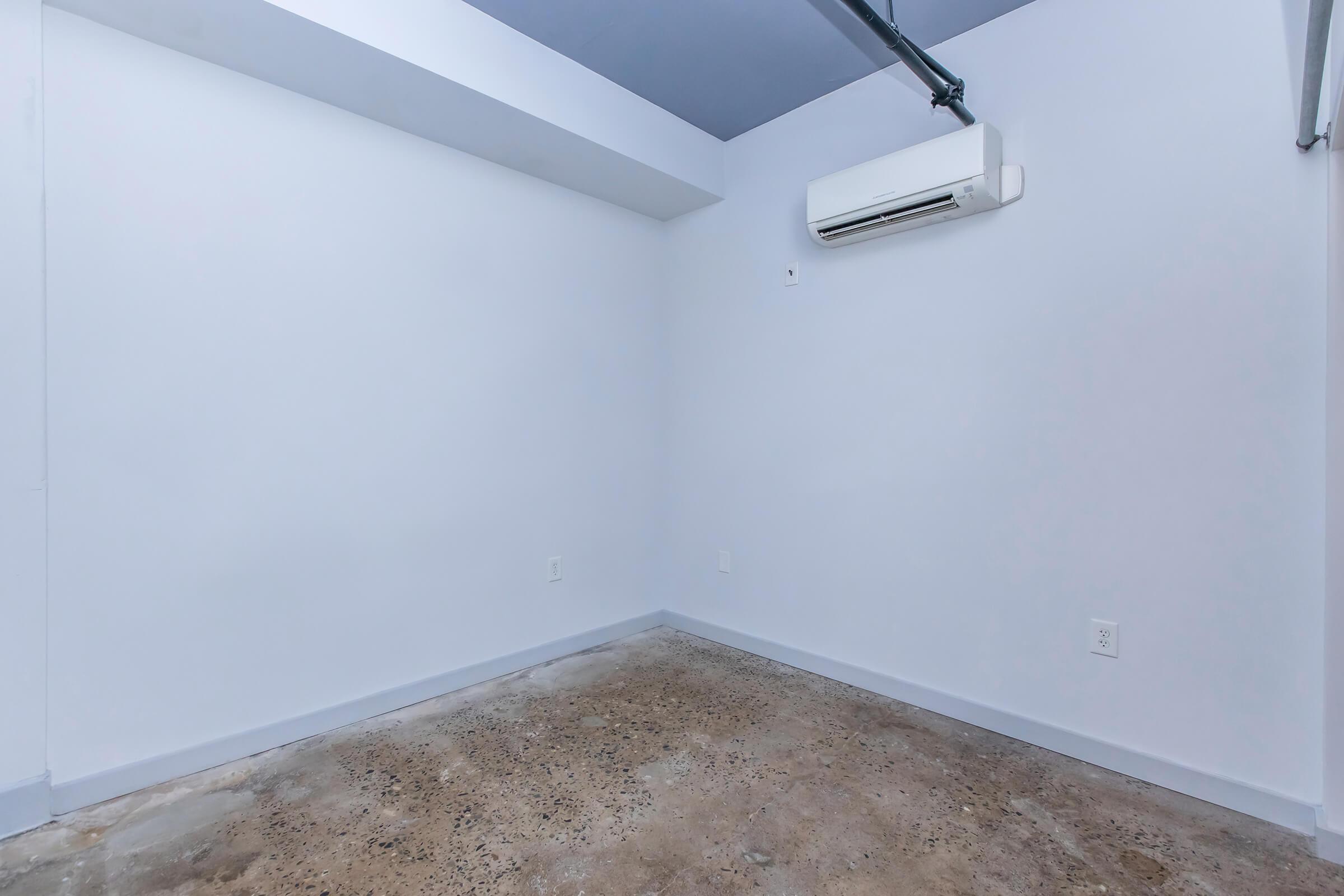 Empty room with light blue walls, a concrete floor, and an air conditioning unit mounted on the wall. The space is uncluttered, featuring a simple and minimalistic design, making it suitable for various uses such as an office or storage area. The ceiling is painted blue, contrasting with the light walls.