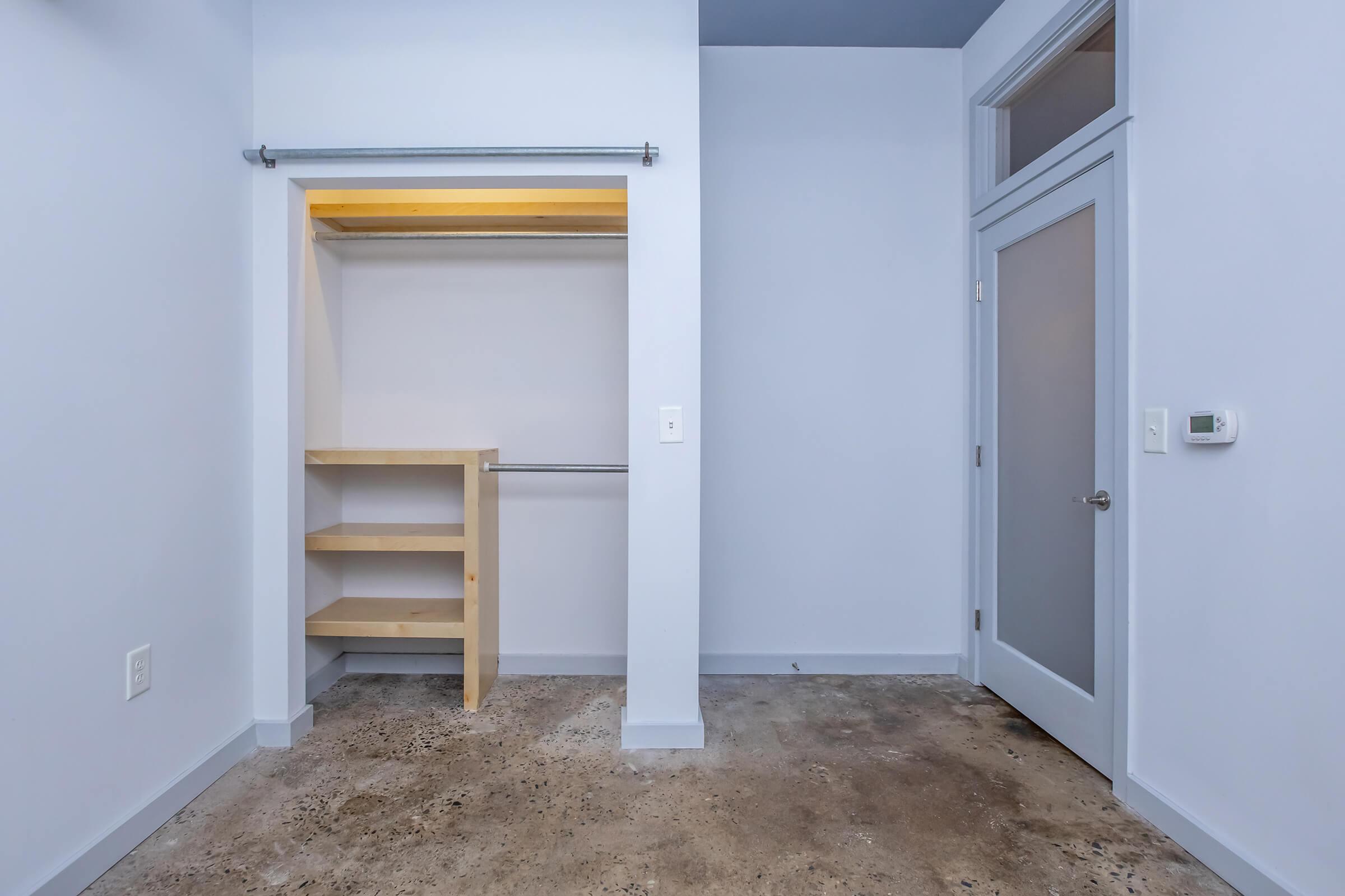 A minimalist room with light blue walls featuring a closet with a wooden shelving unit and a hanging rod. The floor is made of concrete, and there is a door on the right side. Natural light spills into the room, creating a bright and airy atmosphere.
