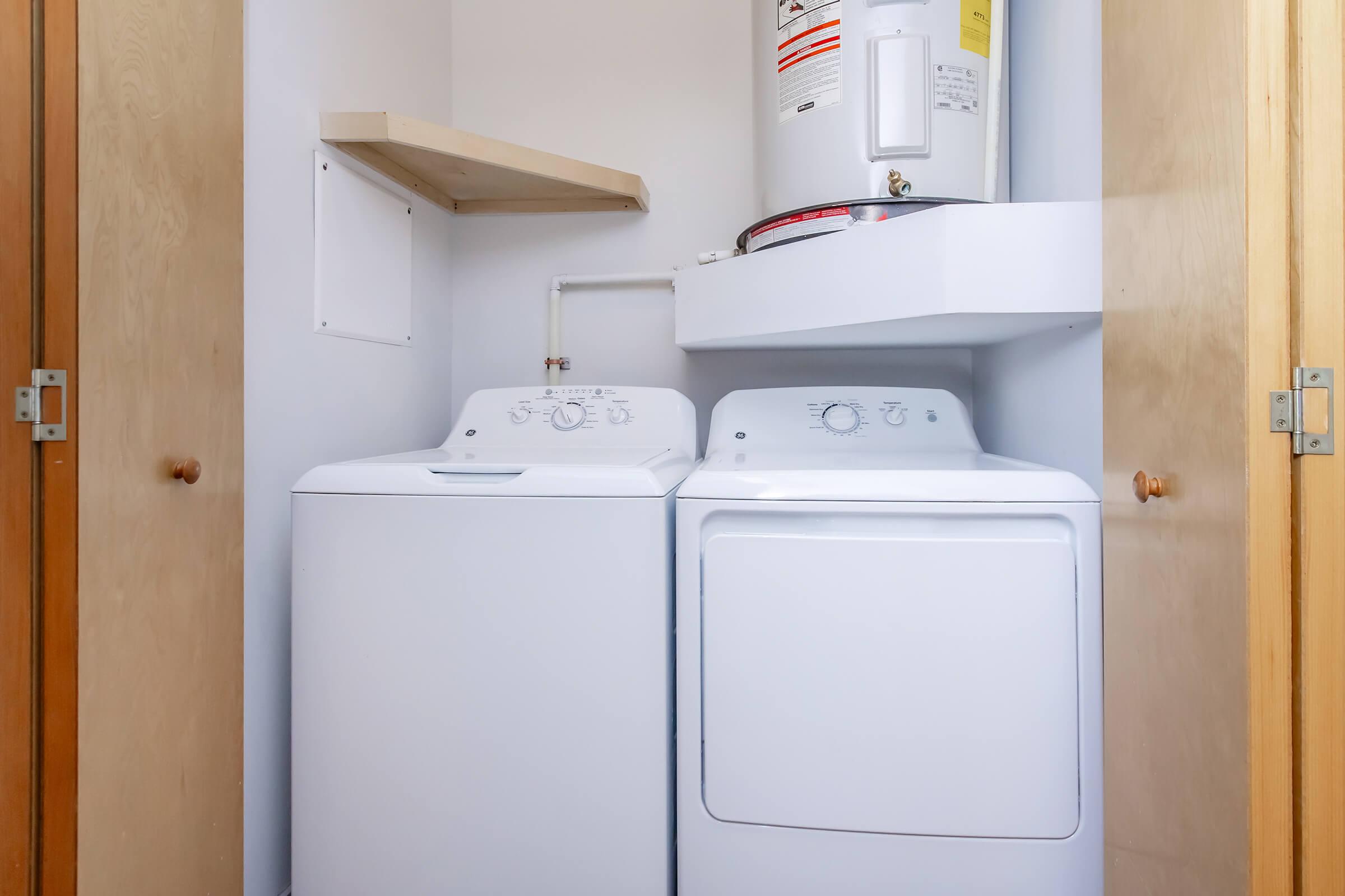 A compact laundry area featuring a white washing machine and dryer stacked side by side. Above the appliances, there is a shelf and an electric water heater mounted on the wall. The overall color scheme is neutral with light wooden doors and a clean, minimalistic design.