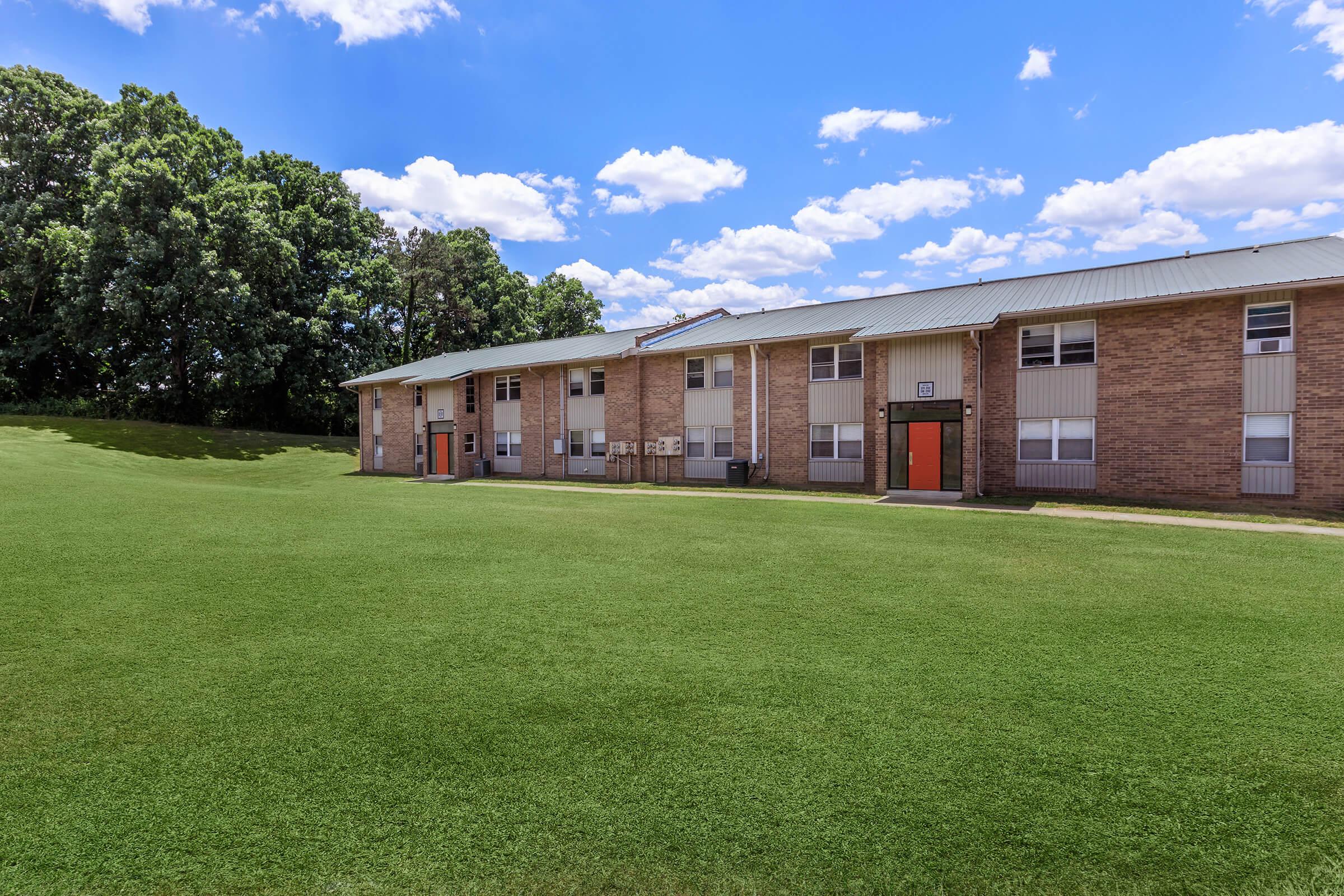 A spacious green lawn in front of a two-story brick building with windows and orange doors, set against a backdrop of a clear blue sky with fluffy white clouds. The area appears well-maintained, providing an inviting atmosphere.