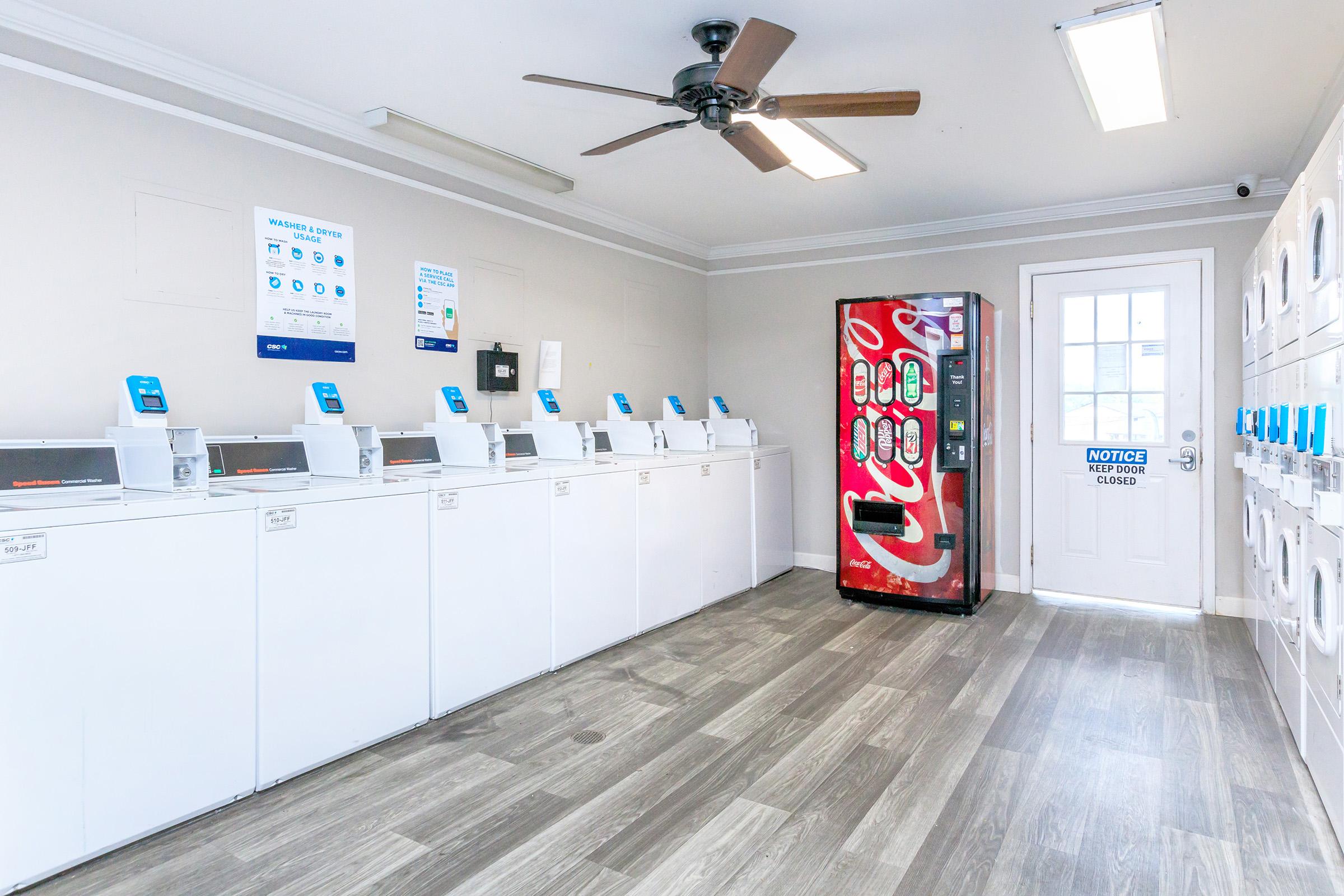 A bright, clean laundromat interior featuring several white washing machines along one wall, a snack vending machine in the corner, and a door with a notice. The room has wooden-style flooring, a ceiling fan, and bright overhead lights, creating a welcoming atmosphere for customers.