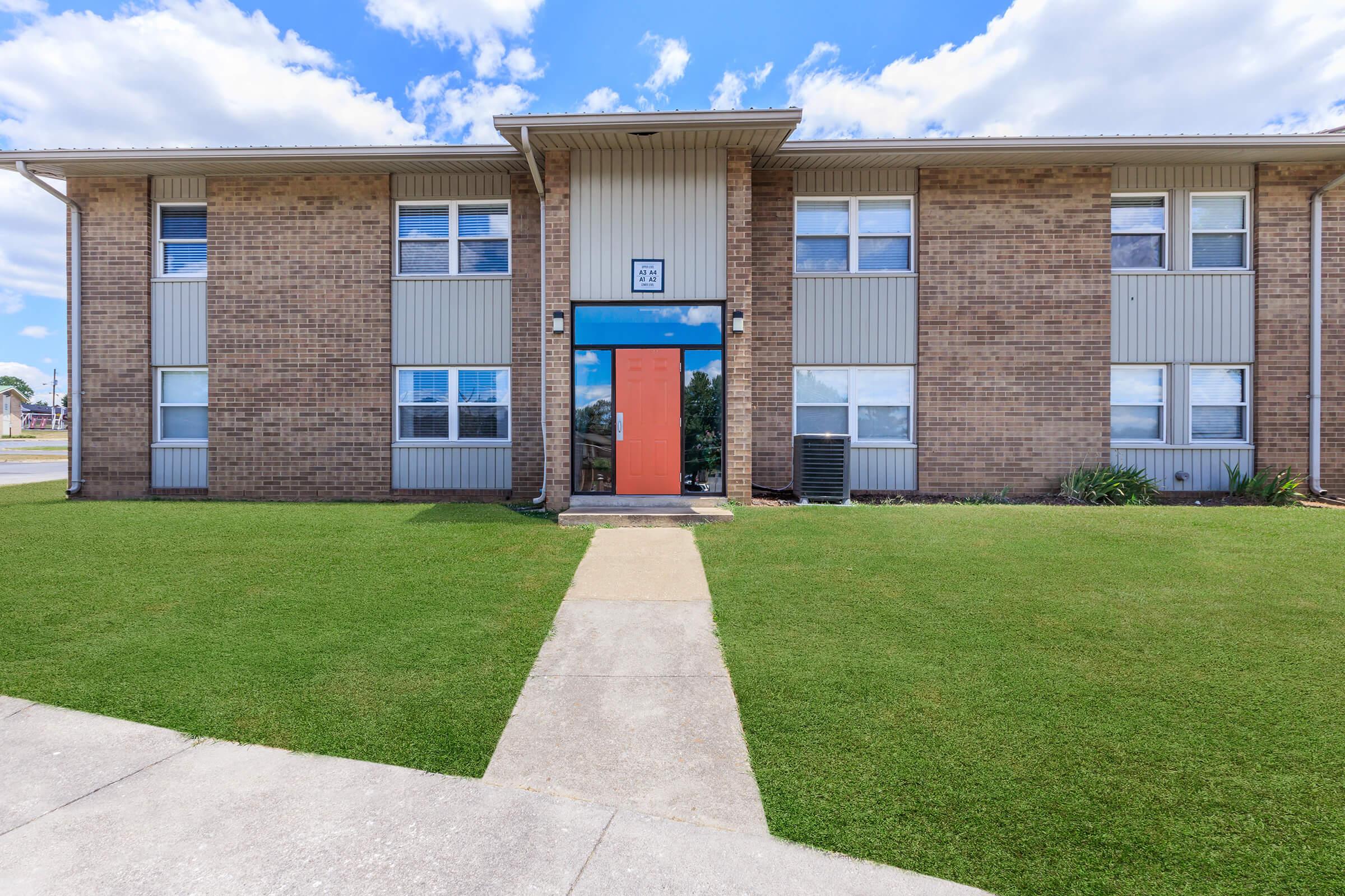 A two-story brick apartment building with a central entrance featuring orange doors. The building has large windows and is surrounded by neatly landscaped green grass. A concrete pathway leads to the entrance, with a clear blue sky and clouds in the background.