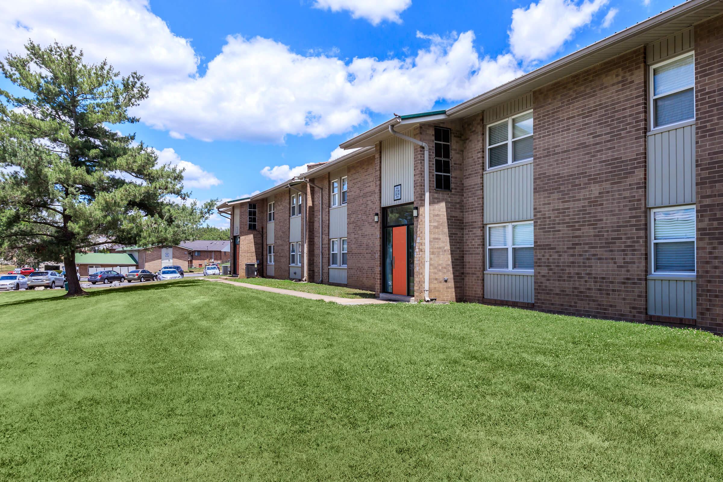 An apartment building featuring multiple stories and large windows, set against a backdrop of blue skies and fluffy white clouds. A well-maintained grassy area surrounds the building, with a tall tree on the left. The building has an orange accent door, creating a welcoming entrance.