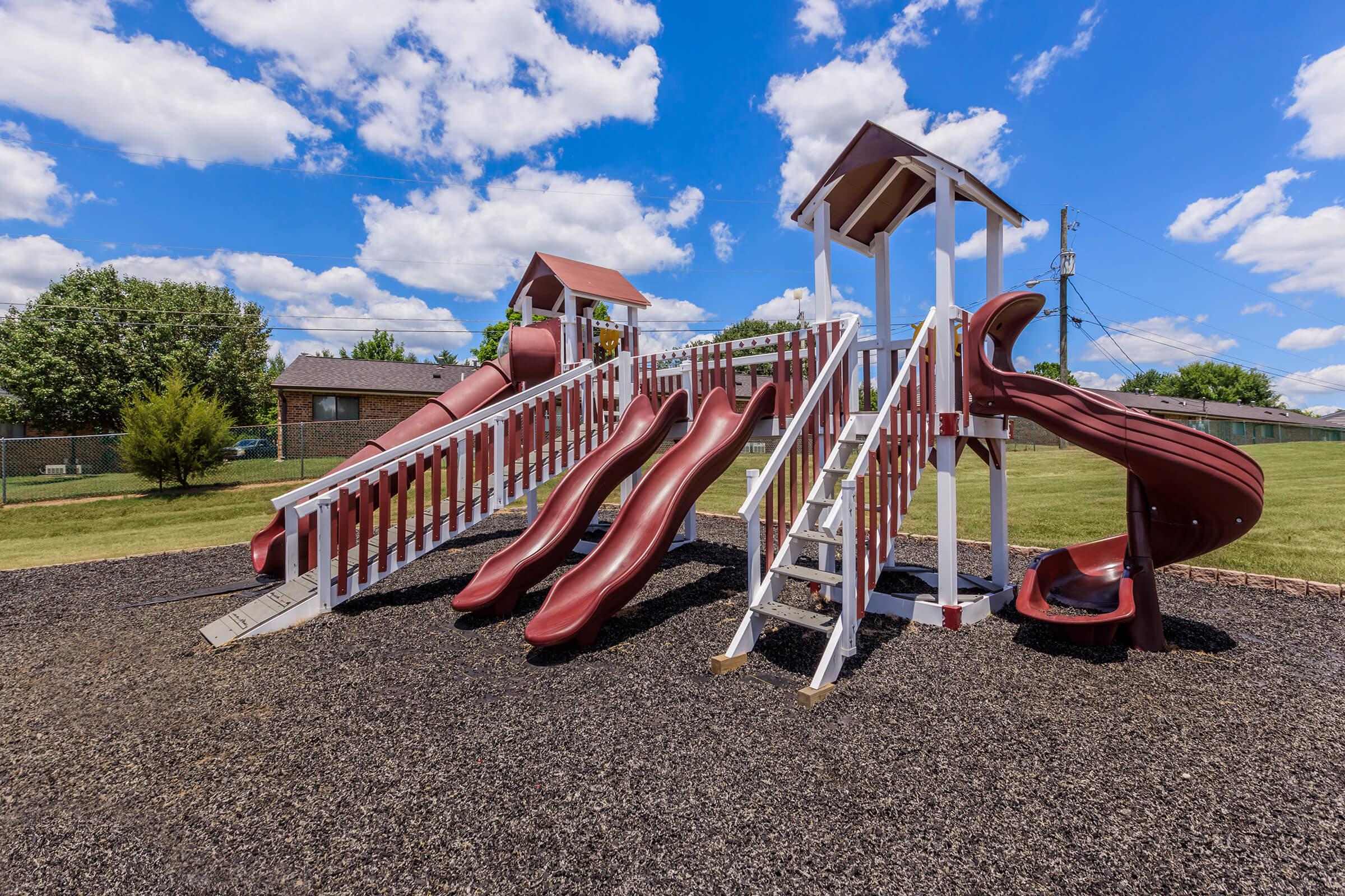 A colorful playground structure featuring multiple red slides, ladders, and a lookout tower, set against a backdrop of green grass and a partly cloudy blue sky. The ground is covered in dark mulch, providing safety for children playing on the equipment.