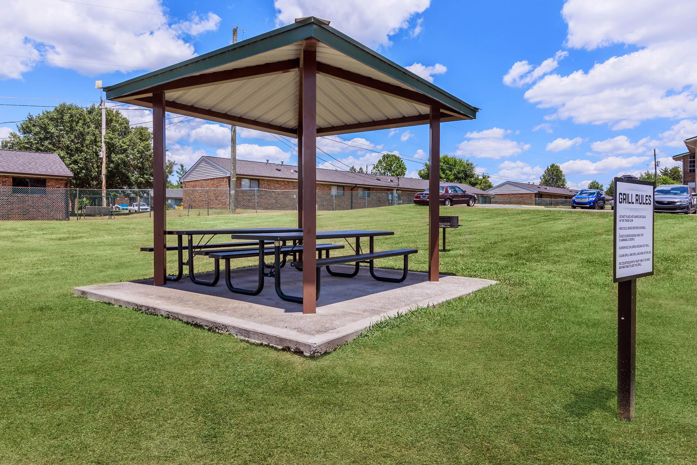 A picnic shelter with a roof and multiple tables is situated on a grassy area. In the background, there are residential buildings and cars parked. A sign nearby outlines grill usage rules. The sky is blue with scattered clouds.