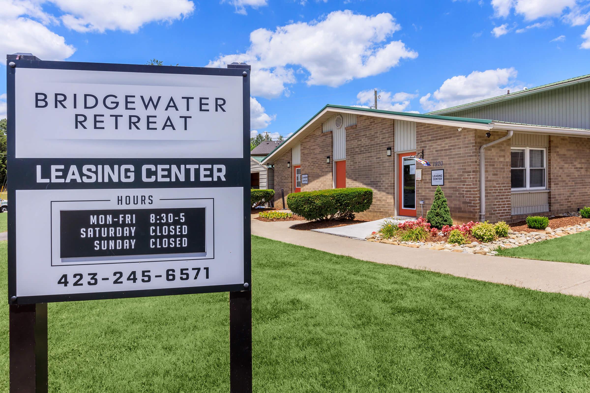 Sign for Bridgewater Retreat Leasing Center displaying operating hours: Monday to Friday from 8:30 AM to 5 PM, closed on Saturday and Sunday. The building is surrounded by well-maintained landscaping and a clear blue sky is visible.