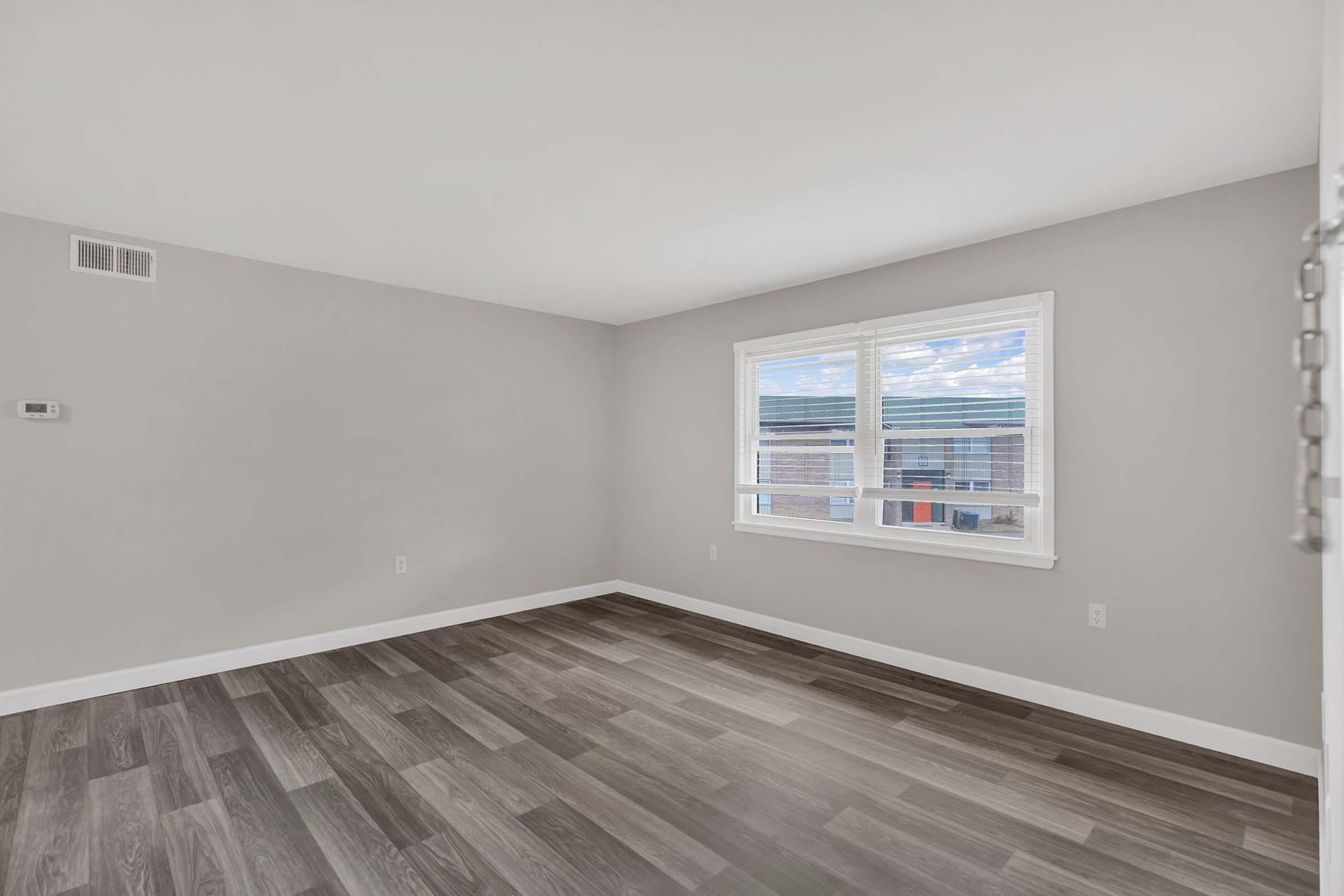 A spacious, empty room with light gray walls and dark wood-like flooring. There is a window with white blinds allowing natural light in, and the room has a simple, modern design. The overall feel is clean and airy, suitable for various uses like a living area or office.