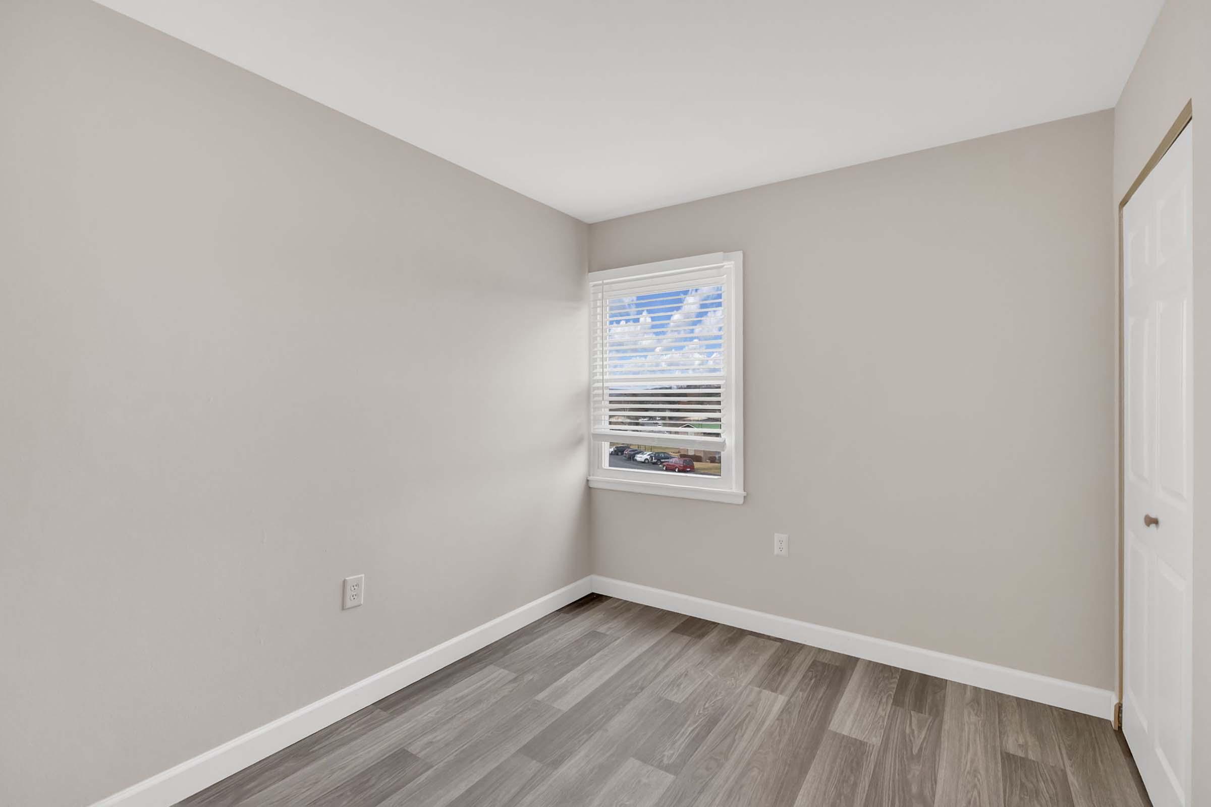 Empty interior room with light gray walls and hardwood-style flooring. A window with white blinds allows natural light to enter, revealing a partly cloudy sky. The room has a simple, clean design with a white door on one side, creating a minimalistic and spacious feel.