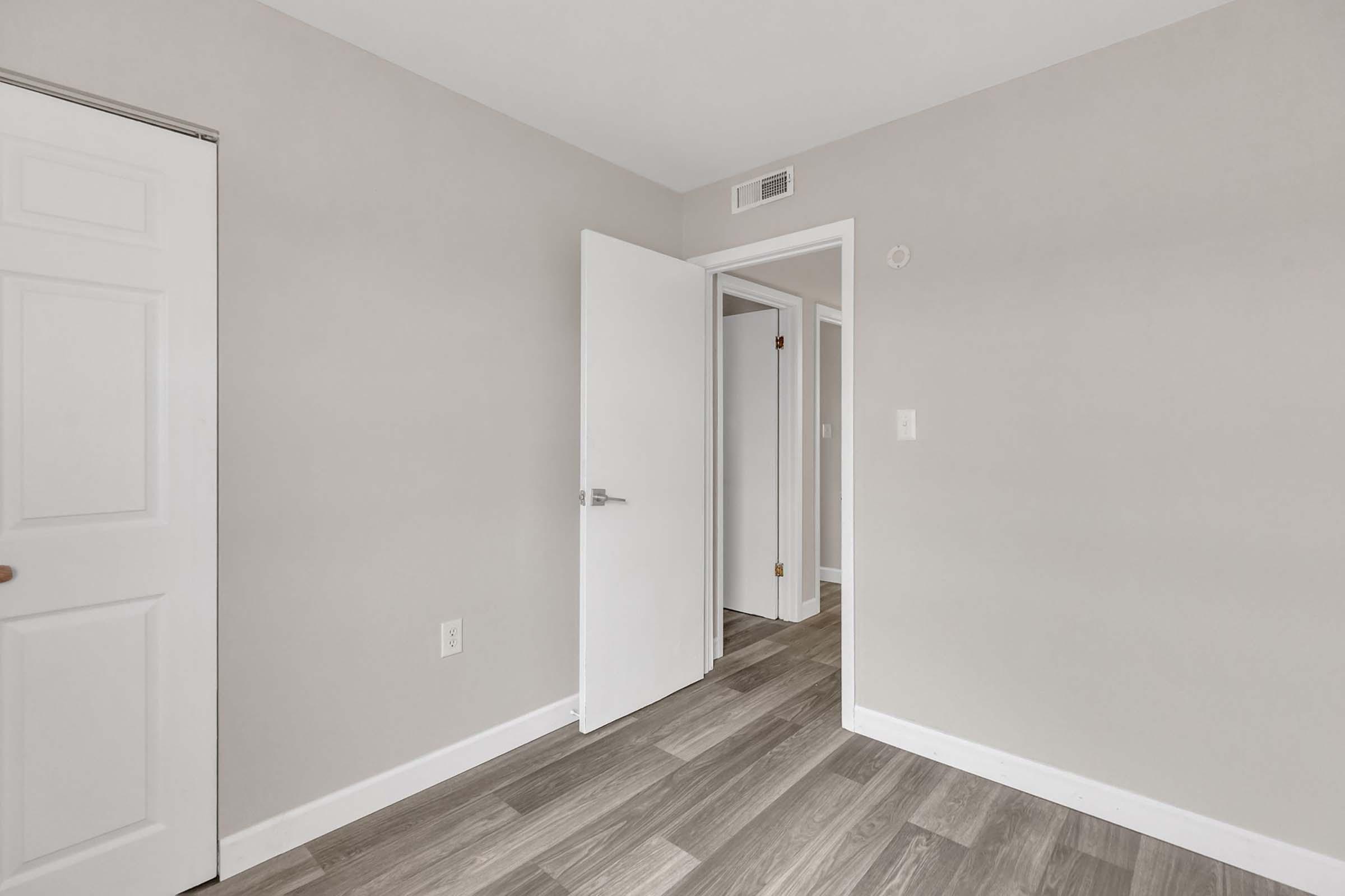 A light-colored room with gray walls and wooden flooring. There is an open white door leading to another room, and a closed white door on the left. The room is well-lit with neutral tones, creating a spacious, clean appearance.