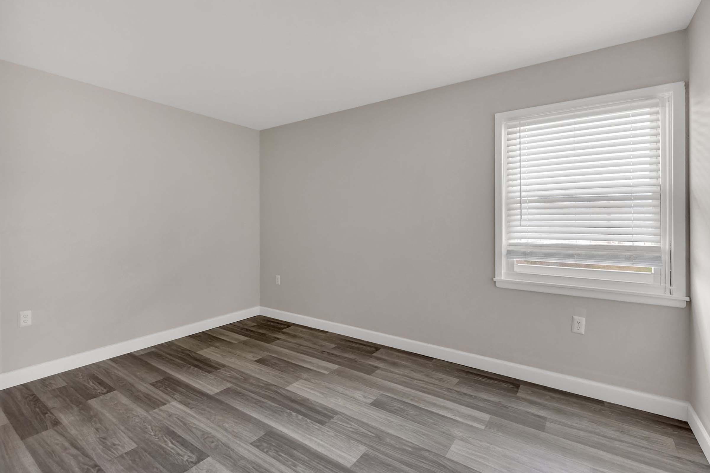 A light gray empty room with hardwood flooring, featuring a white window with blinds and plain walls. The space appears clean and minimalistic, emphasizing its emptiness and potential for decoration or furniture placement.