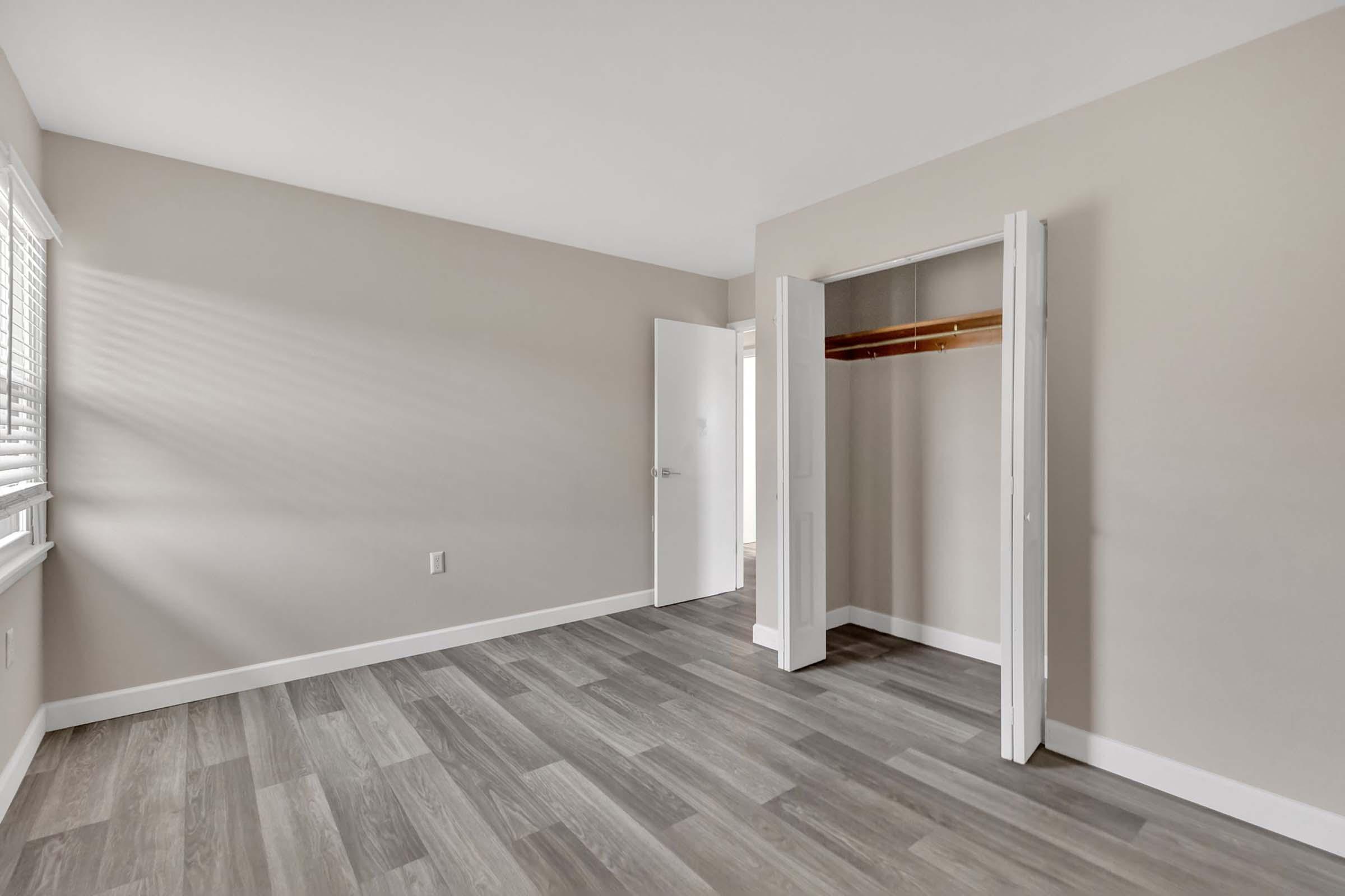 A spacious, empty room with light gray walls and wooden flooring. Natural light streams in through a window with blinds. There is a closed door leading to another area and an open closet with a wooden shelf on one side, contributing to the minimalist and clean aesthetic.