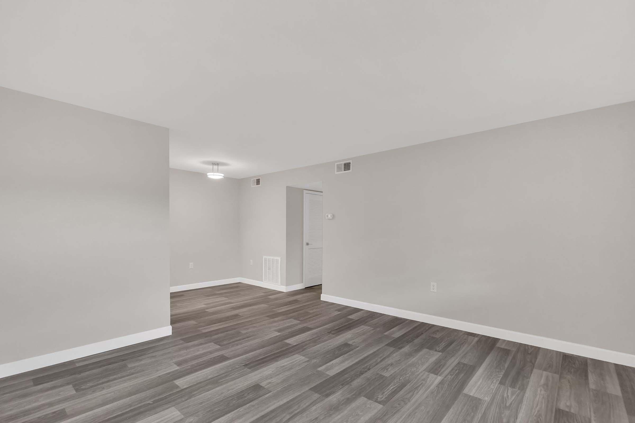 A spacious, empty room with light gray walls and a light wood-like floor. A circular ceiling light provides illumination. An open doorway leads to another room, and a small wall vent is visible. The overall ambiance is clean and modern, suggesting a fresh living space ready for decoration and furniture.