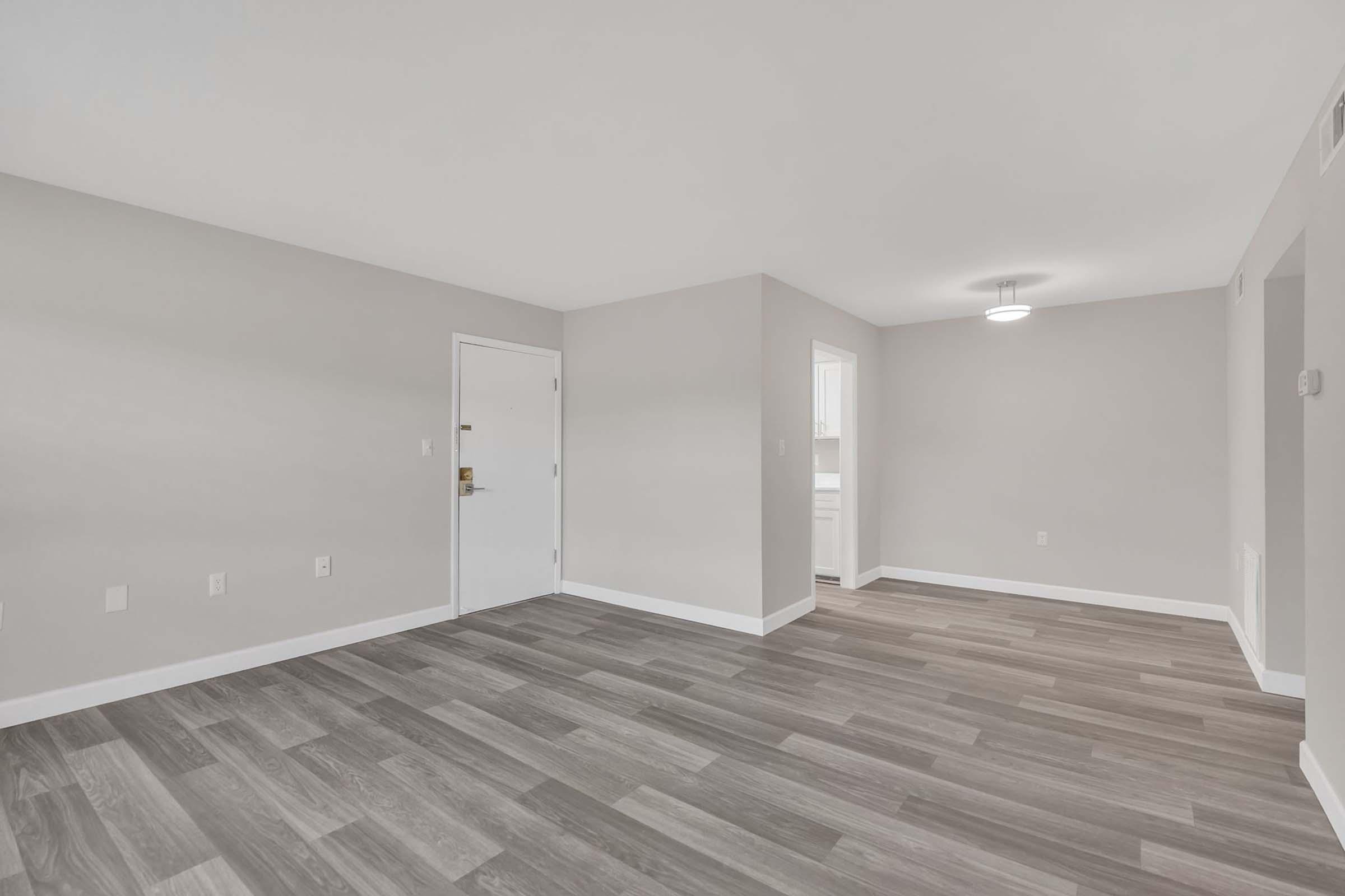 A spacious, empty room with light gray walls and a modern, neutral-toned hardwood floor. A doorway leads to another room, and a light fixture hangs from the ceiling. Natural light enters through a window, enhancing the bright and clean atmosphere.