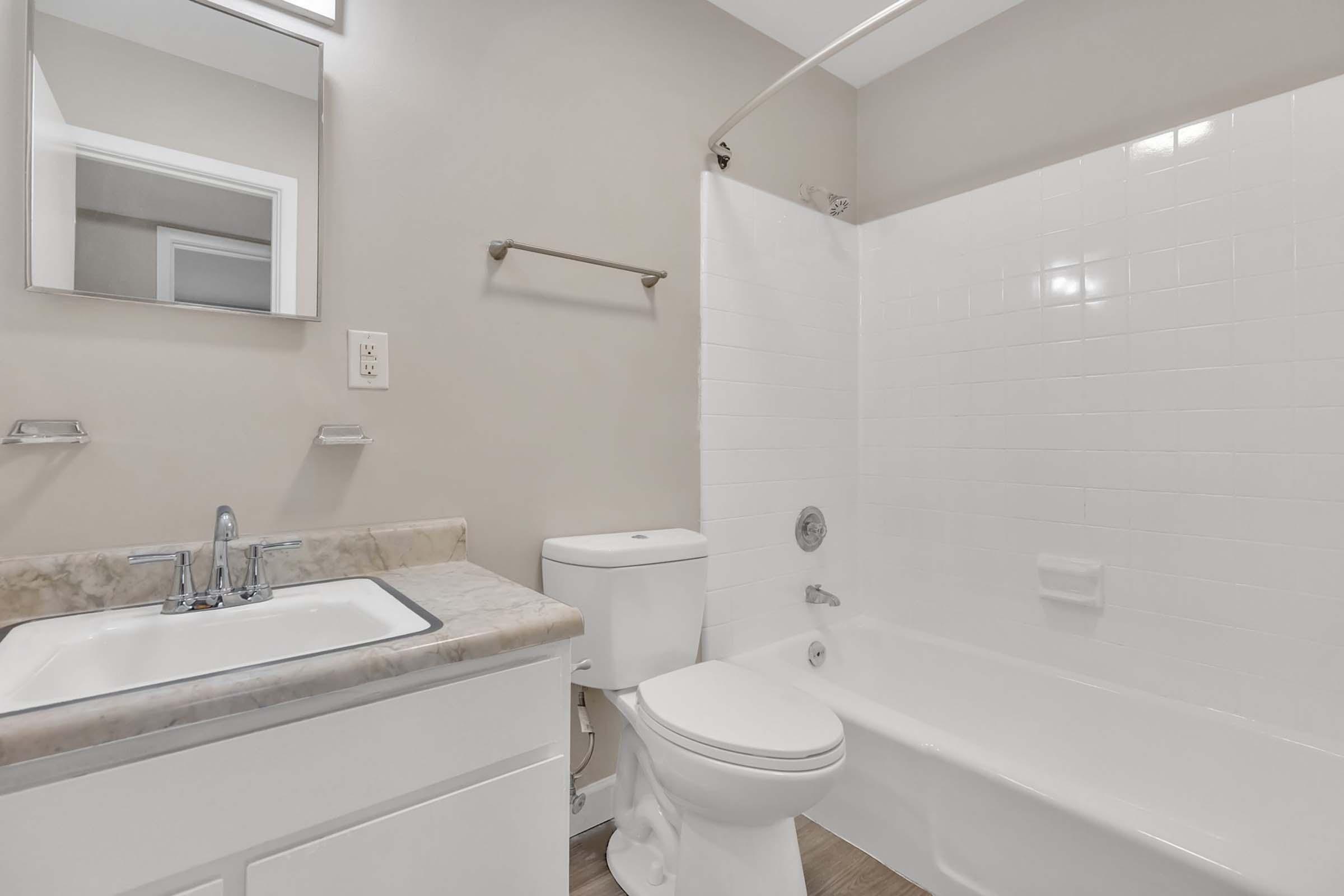 A clean and modern bathroom featuring a white bathtub with a shower, a rectangular mirror above a sink with a single faucet, and a toilet nearby. The walls are painted in a soft beige color, and the floor is a light wood tone, creating a bright and inviting atmosphere.