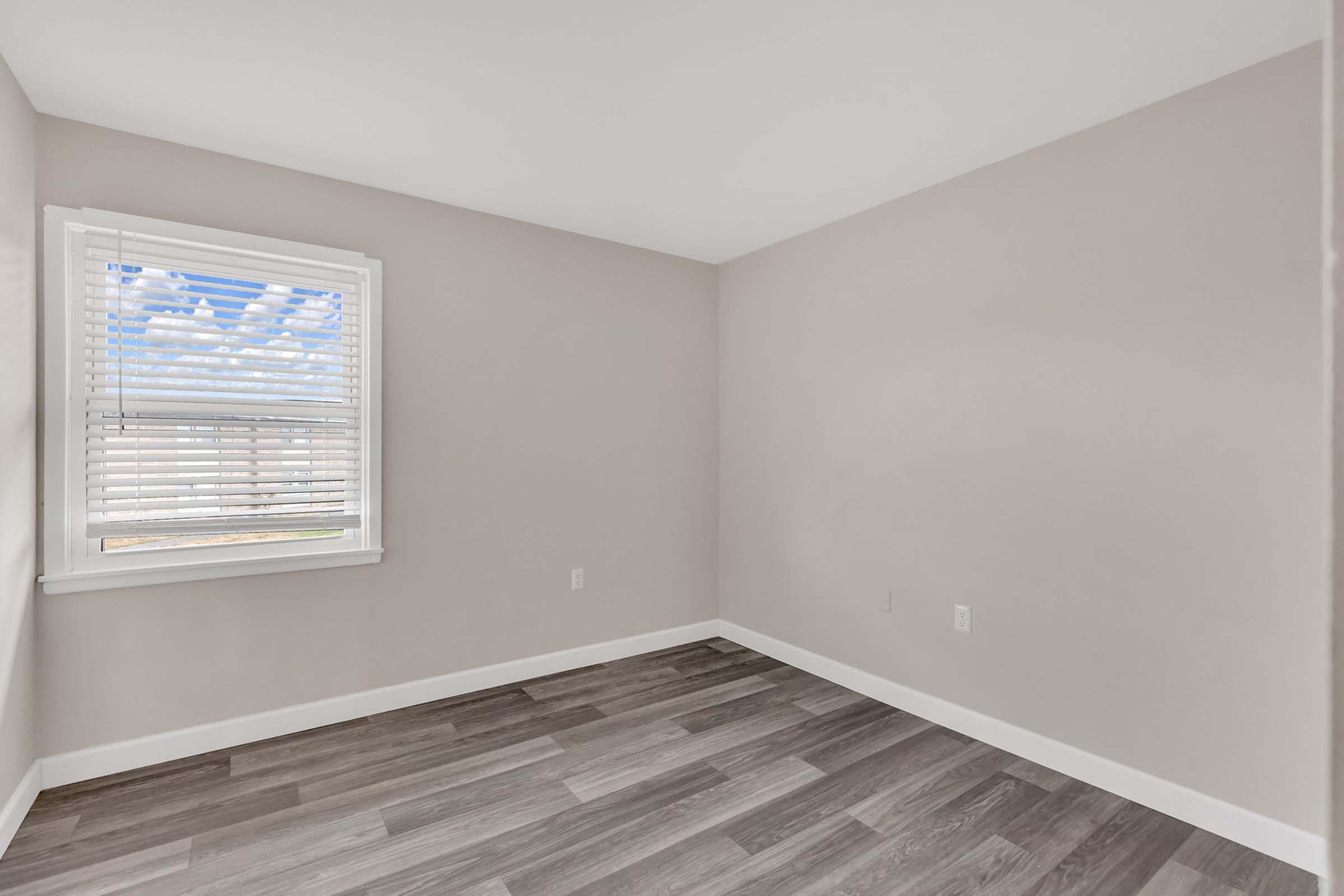 A spacious, empty room featuring light gray walls, a large window with white blinds allowing natural light in, and a wood-look floor. The room has a minimalist design and clean lines, creating a bright and airy atmosphere.