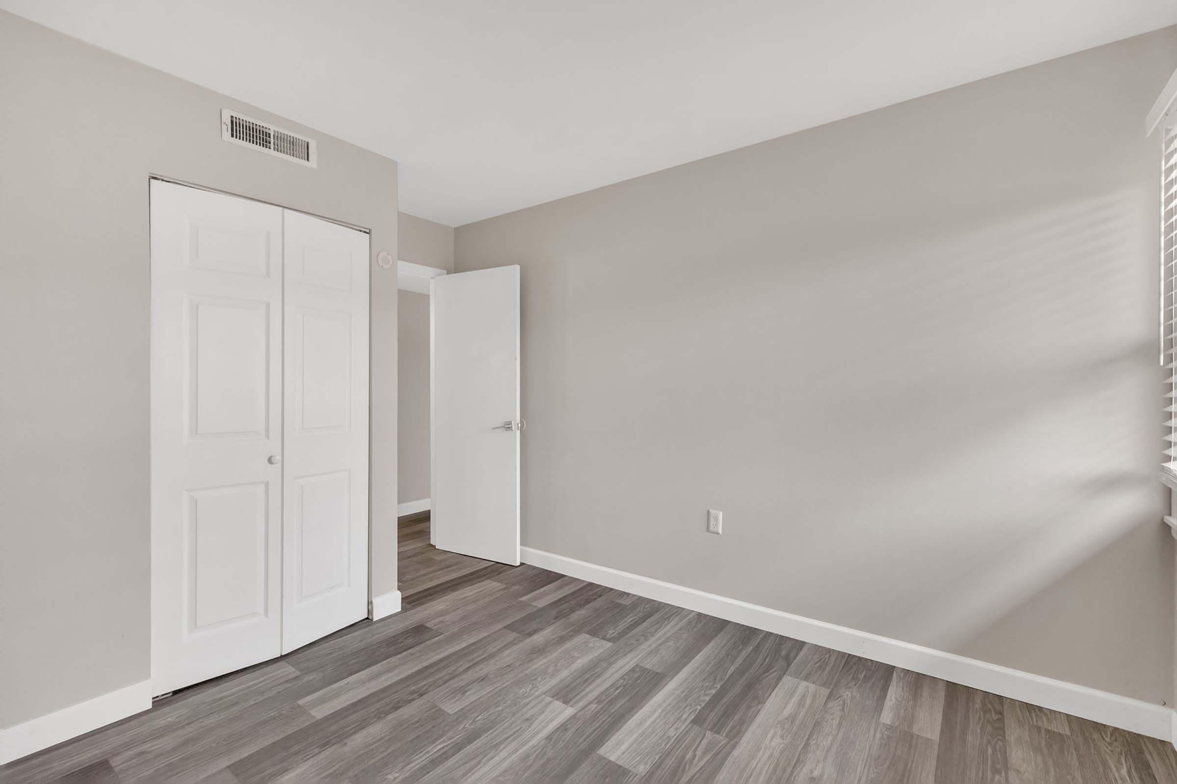 A minimalistic room featuring light gray walls, a floor of wooden planks, and two doors—one with double panels and another single door. Natural light filters in through a window, highlighting the simplicity of the space.