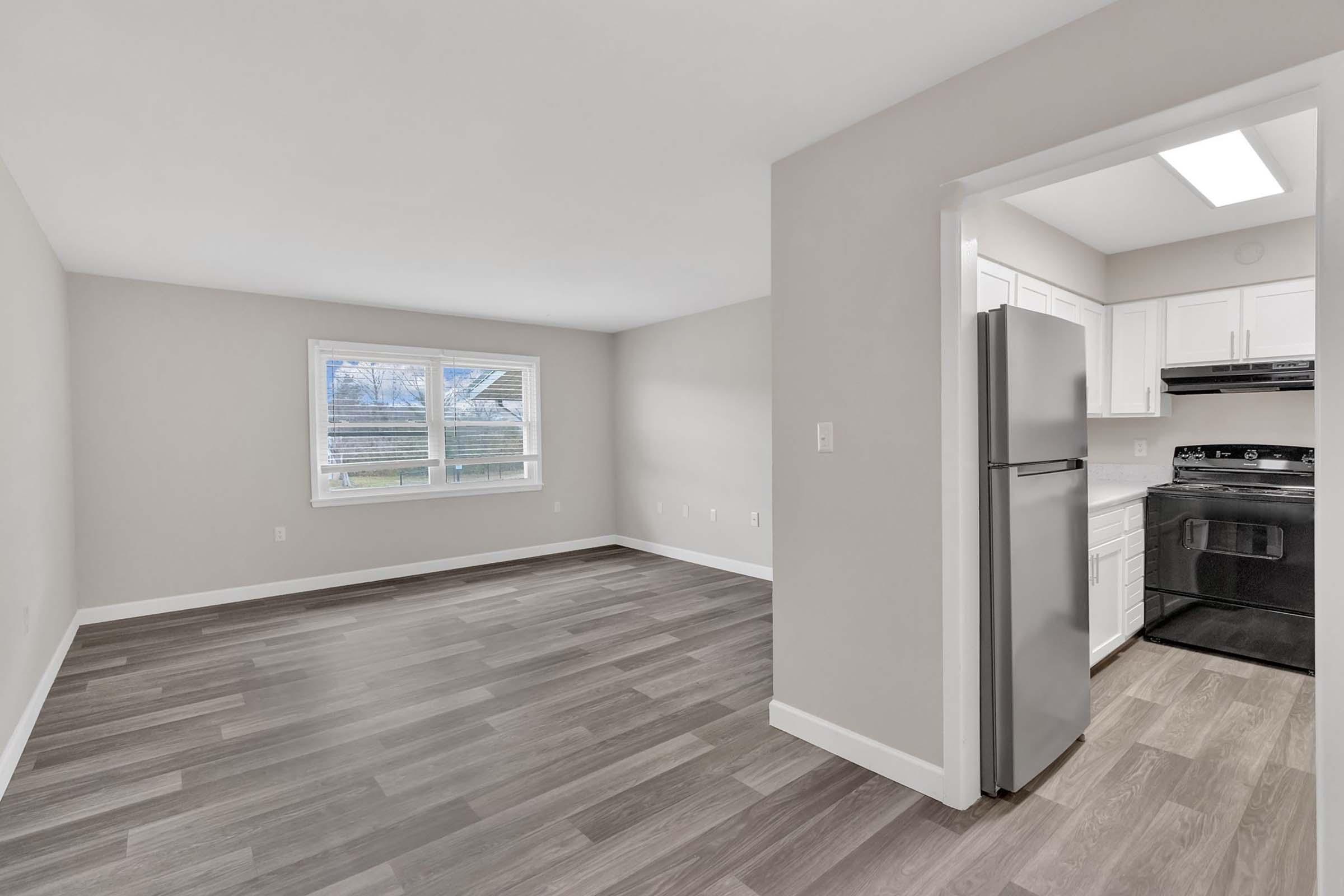 A bright, empty living space featuring light gray walls and new flooring. There is a window with natural light illuminating the room. A kitchen area is visible on the right, showcasing white cabinetry, a refrigerator, and a black stove. The layout is open and modern, suitable for various decor styles.