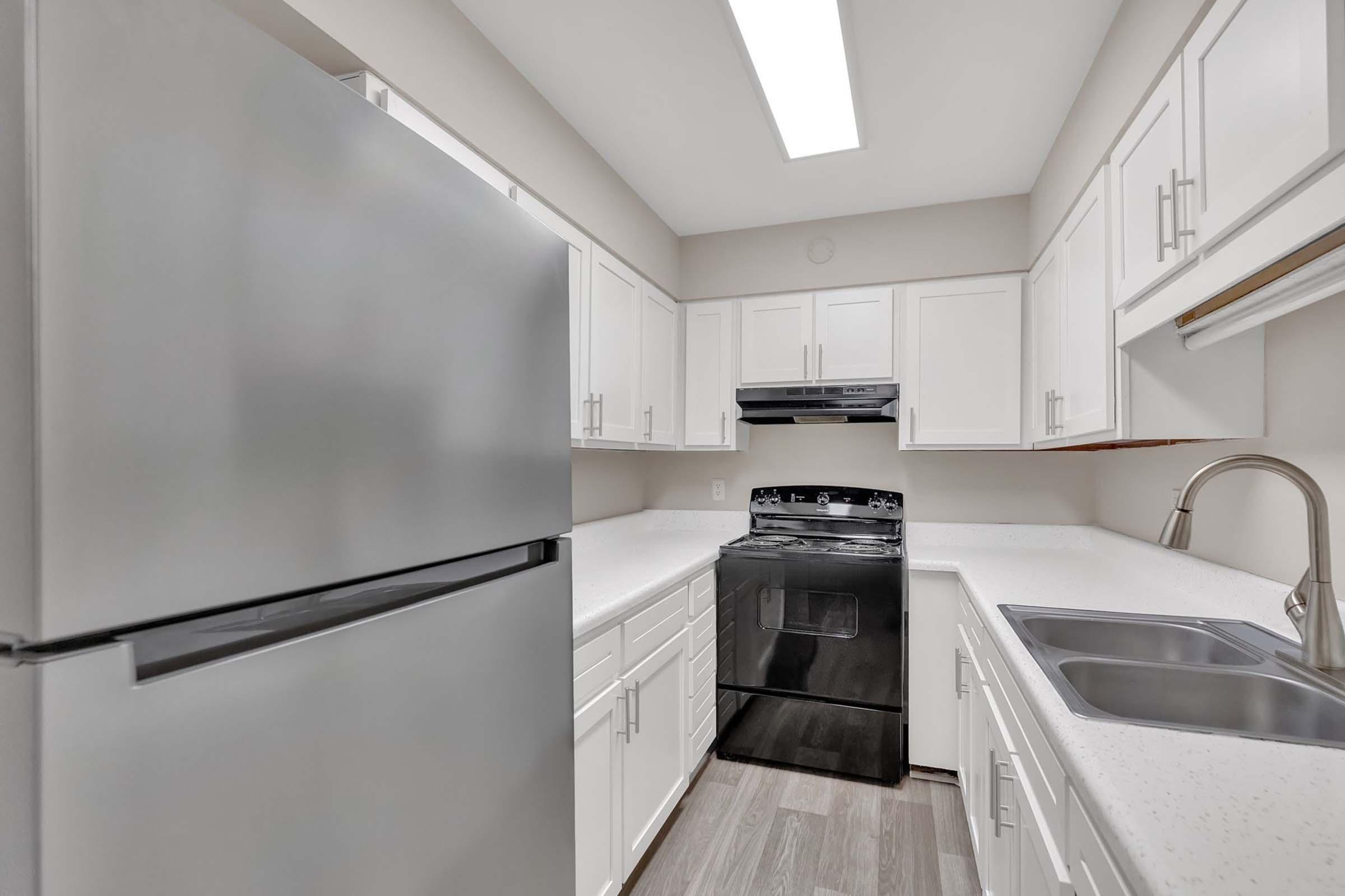 A modern kitchen featuring a stainless steel refrigerator, a black gas stove, and a double sink. The cabinets are white, and the countertop is light-colored, creating a clean and spacious atmosphere. Natural light comes from a ceiling-mounted fixture, illuminating the room.