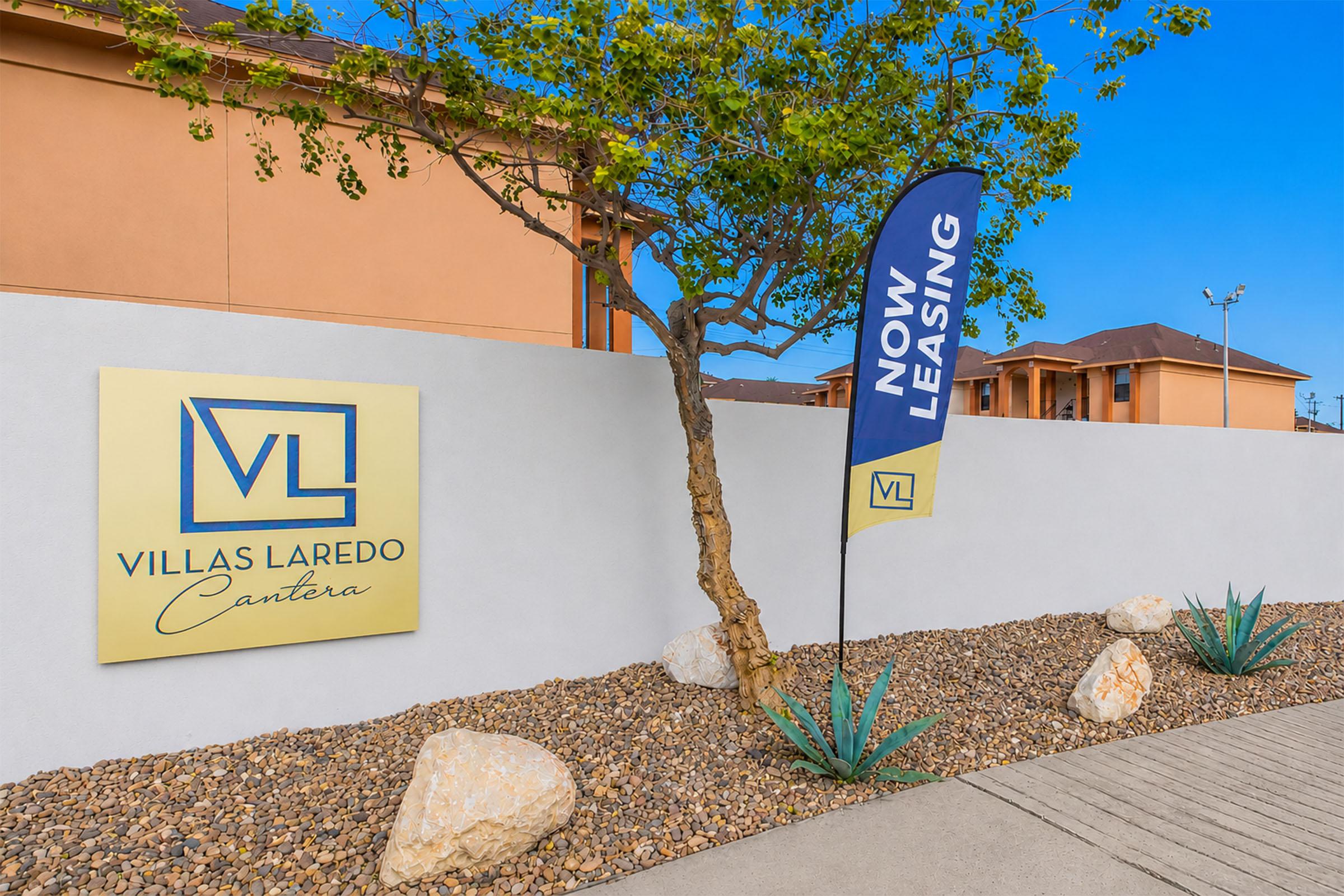 Signage for Villas Laredo Cantera, featuring a large logo with "VL" and the text "Now Leasing" on a flag. A landscaped area with a few rocks and agave plants is in front of a white wall, with buildings visible in the background against a clear blue sky.