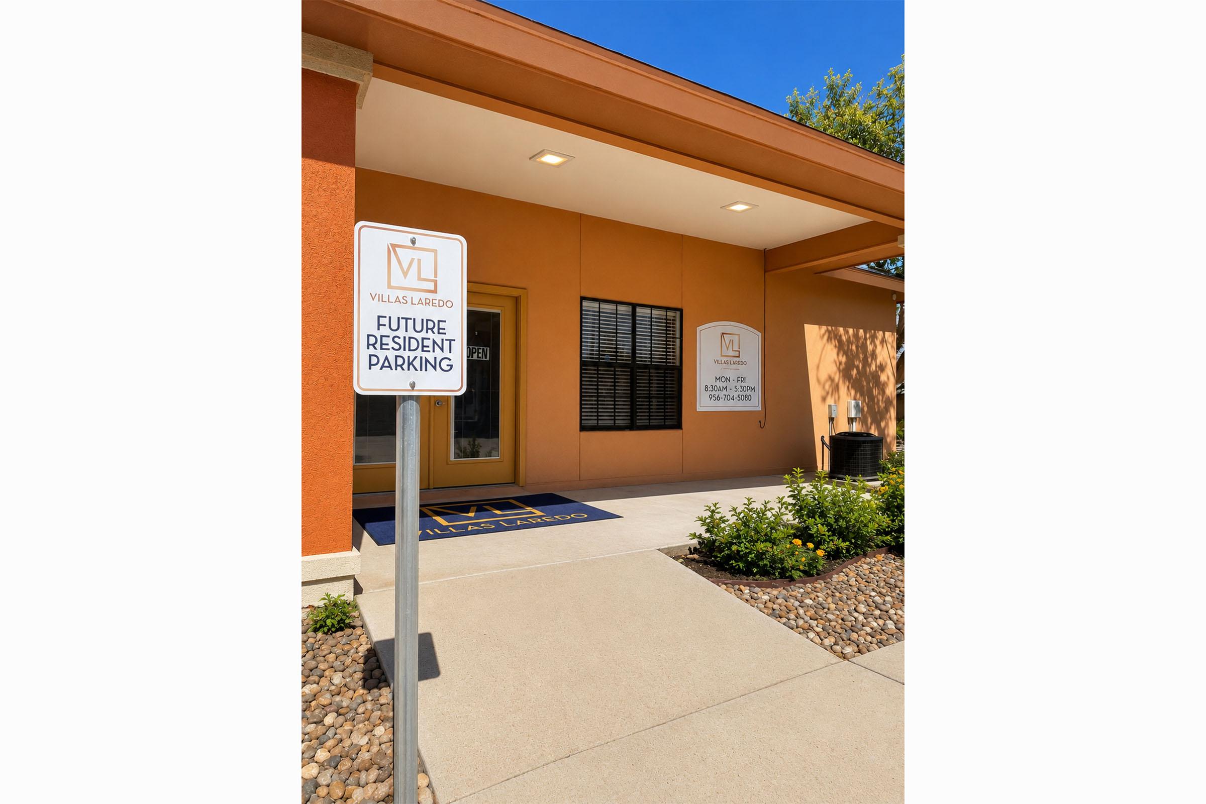 An exterior view of a building with a sign indicating "Future Resident Parking" next to a decorative entryway. The building features orange walls, large glass doors, and a small landscaped area with rocks and plants. Clear blue sky overhead enhances the bright atmosphere.