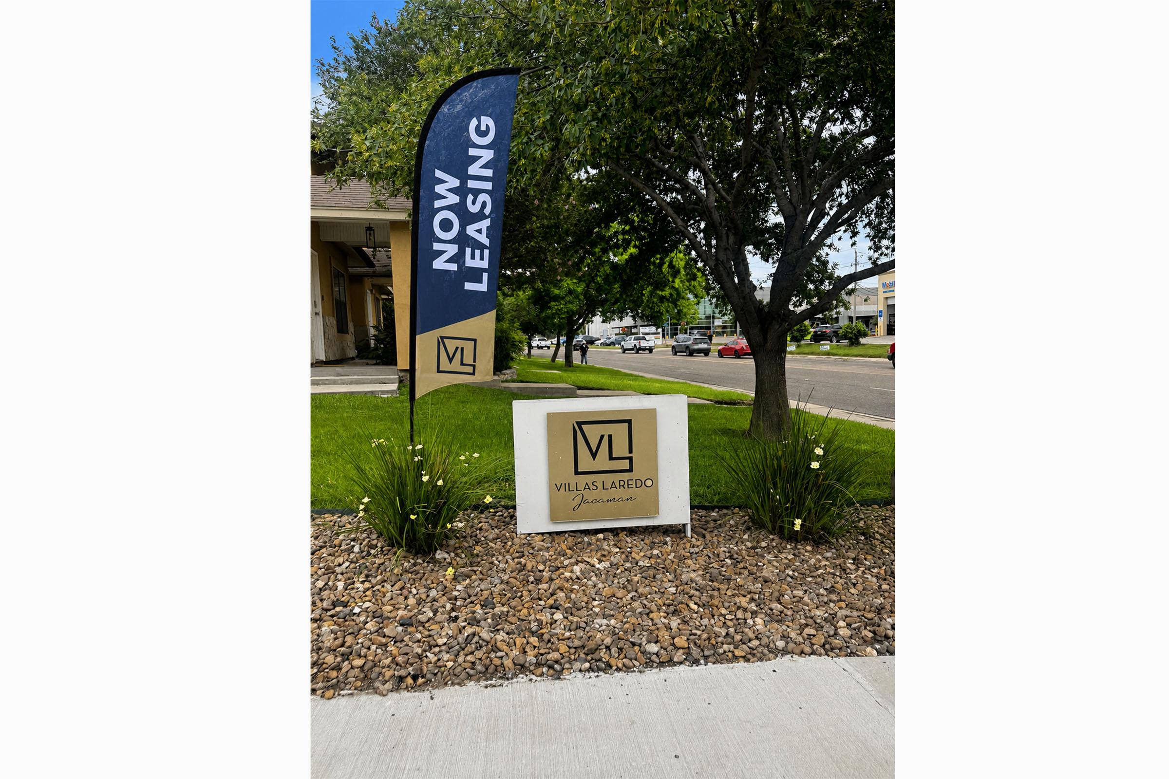 Sign for "Villas Laredo" indicating that apartments are available for lease. The sign features a prominent "Now Leasing" banner, surrounded by landscaping with shrubs and flowers, set against a sunny, blue sky.