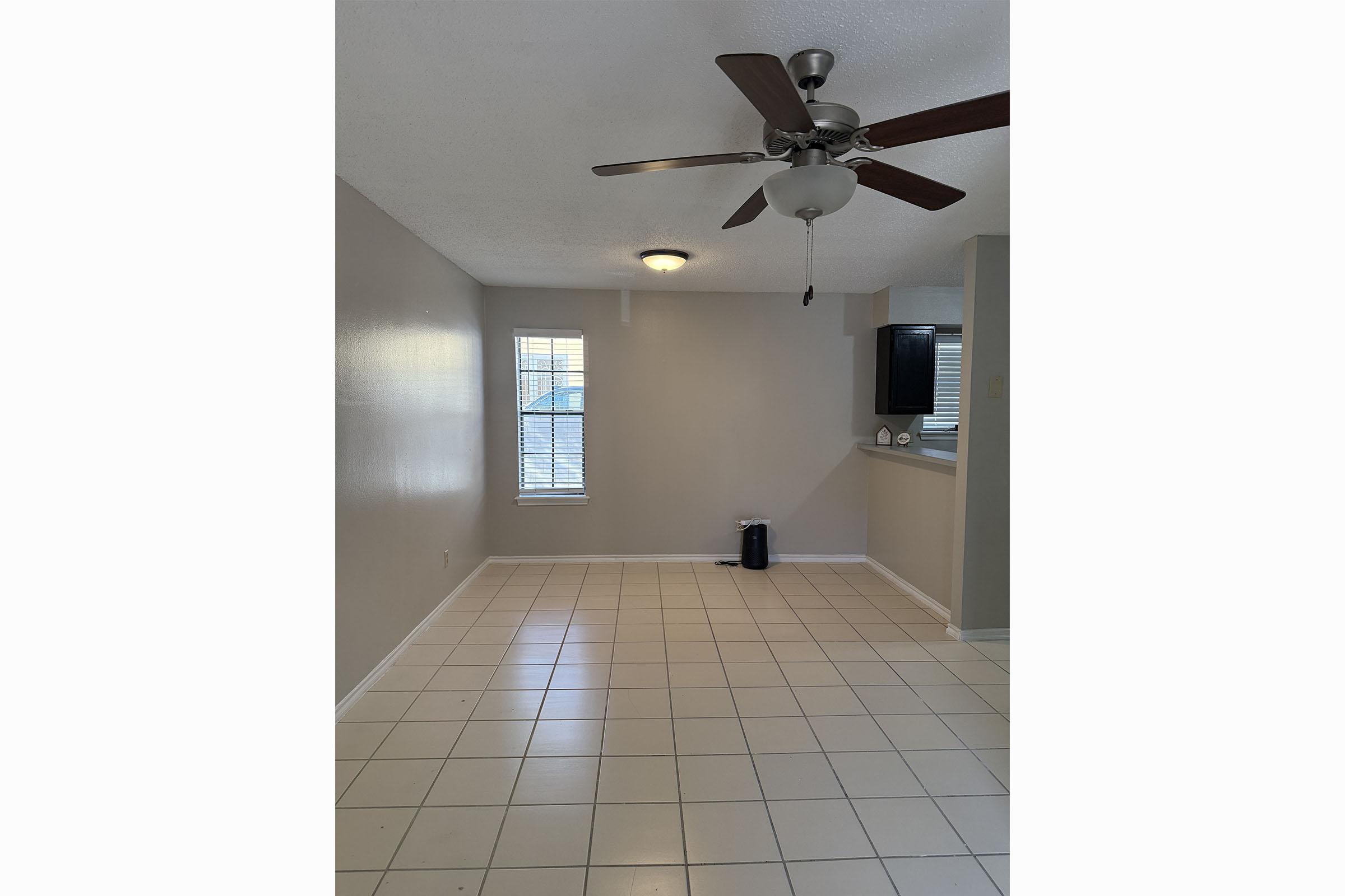 An empty room with light-colored walls and tiled flooring. A ceiling fan is mounted above, and there is a window with blinds on one wall. A small black appliance is on the floor, and a kitchen area with a dark countertop is visible in the background. Natural light illuminates the space.
