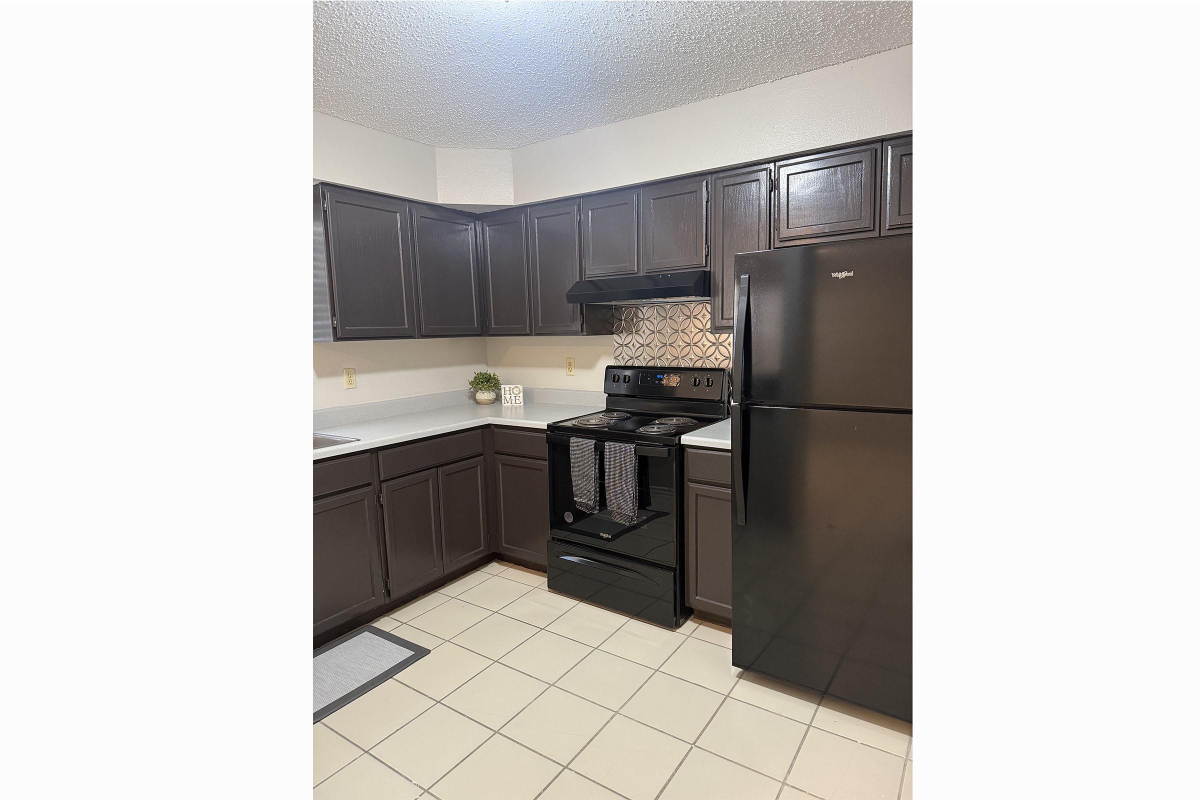 Modern kitchen with dark wood cabinets, a black stove and refrigerator, and light-colored countertops. The backsplash features a decorative tile design. There is a small plant on the counter, and the flooring consists of light-colored tiles. Natural light is coming from a window above the sink.
