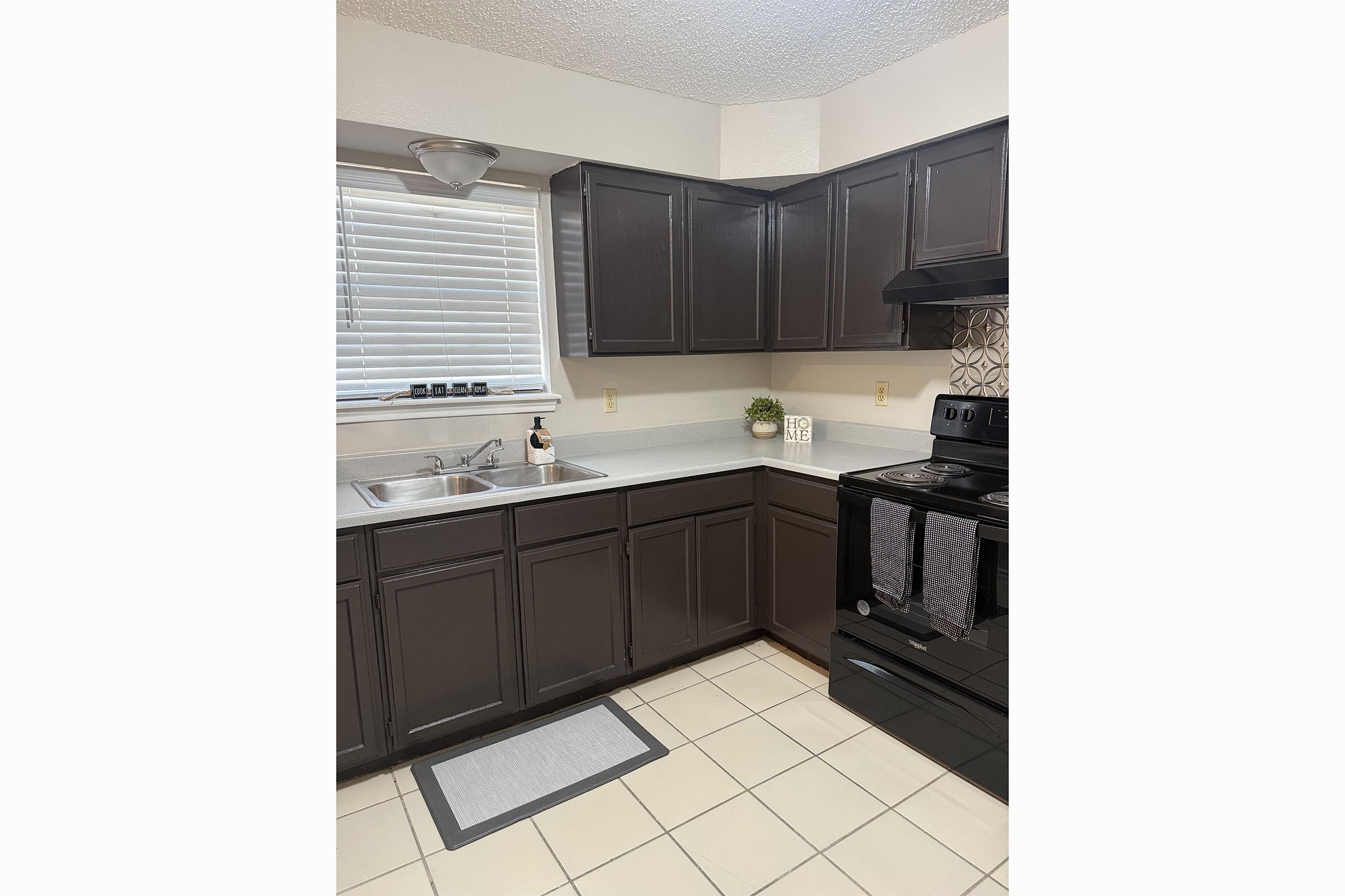 A modern kitchen featuring dark cabinetry, a white countertop, a sink under a window with blinds, and an electric stove. The floor is tiled, and there is a small grey rug in front of the sink. Decorative items include a potted plant on the countertop.