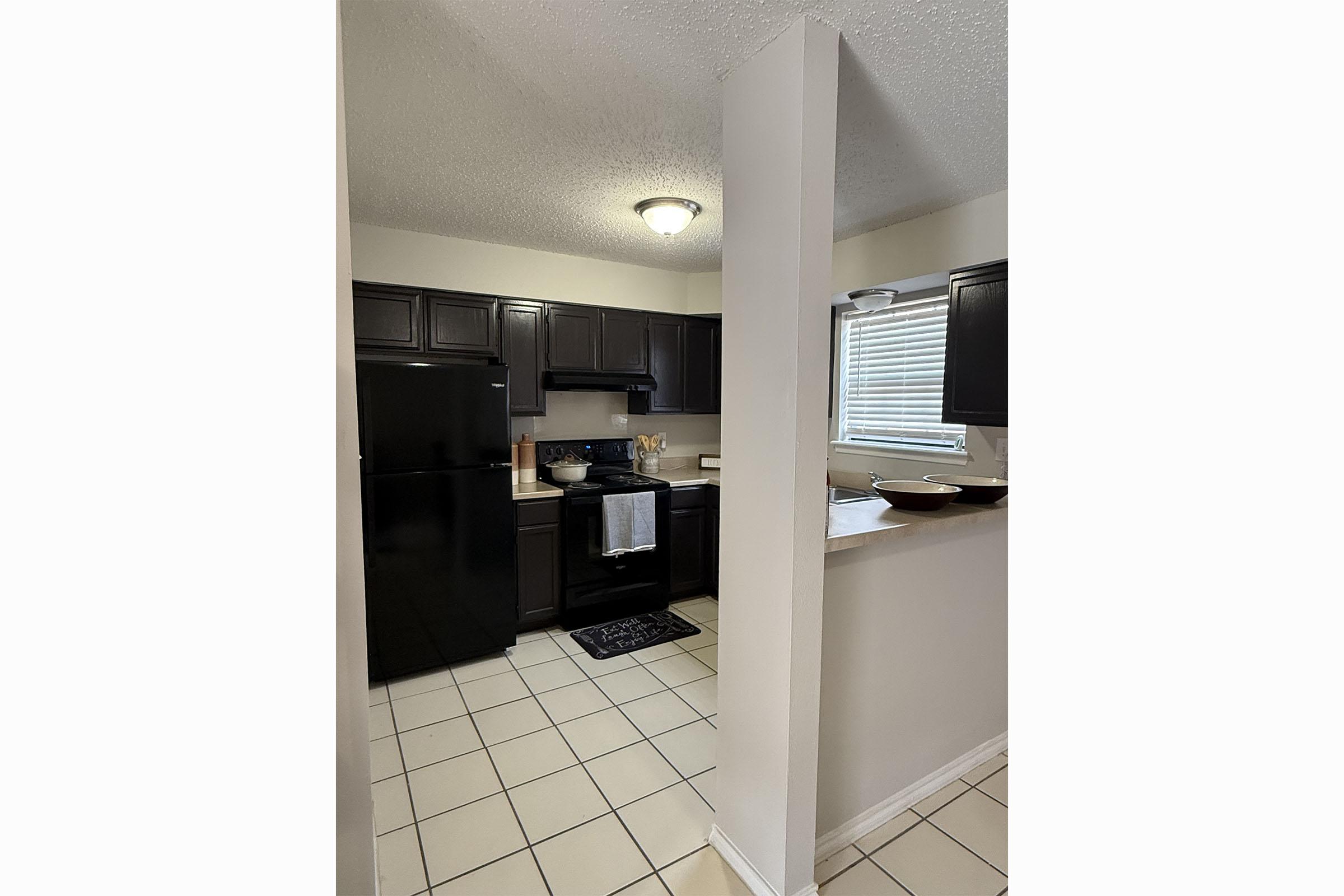 Modern kitchen featuring dark wood cabinets, a black refrigerator, and a black stove. The space has white tiled flooring and a window providing natural light. A countertop with a bowl and a towel hangs from the stove, adding to the functional aesthetic of the room.