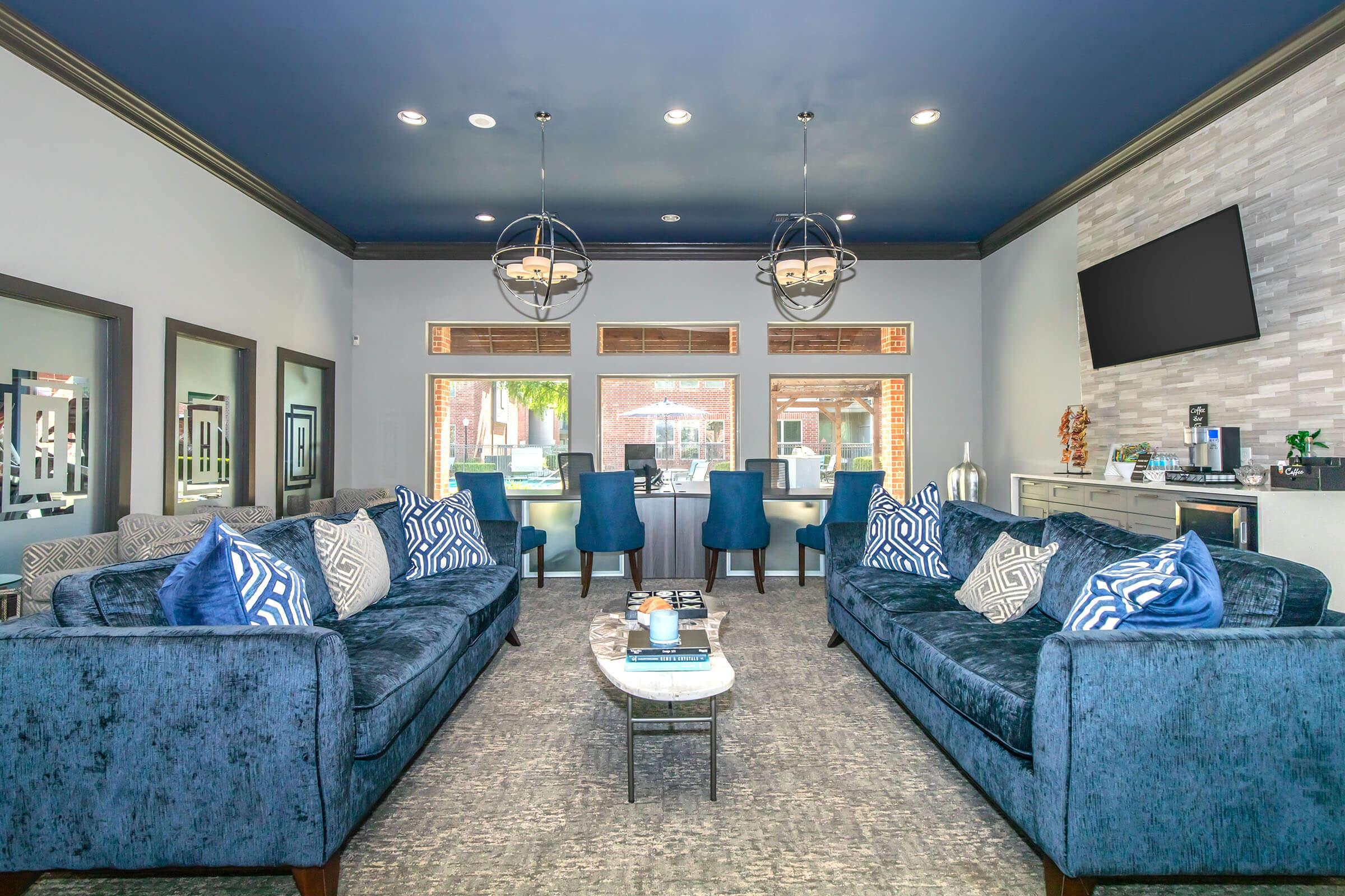 A modern living room featuring two blue velvet sofas with decorative cushions, a sleek coffee table at the center, and large windows allowing natural light. The ceiling is painted dark blue, with stylish light fixtures hanging above. The decor includes mirrors and a flat-screen TV mounted on the wall, creating a cozy yet sophisticated atmosphere.