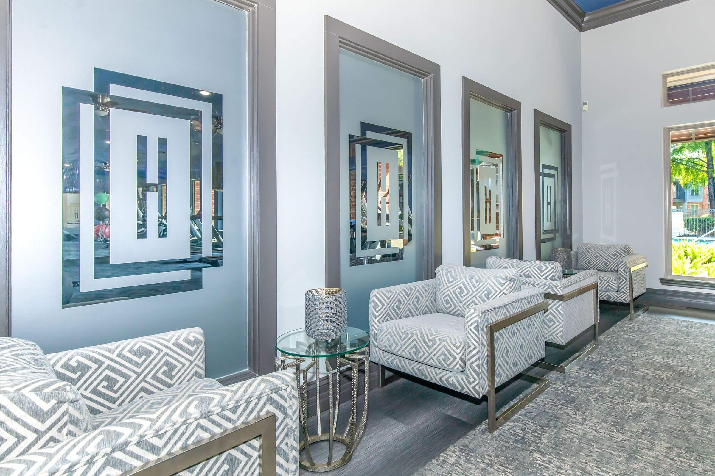 A modern waiting area featuring four patterned chairs with metallic frames, a small glass table beside one chair, and stylish wall mirrors displaying abstract geometric designs. The space is brightly lit and has neutral-colored walls, creating a sophisticated and inviting atmosphere.