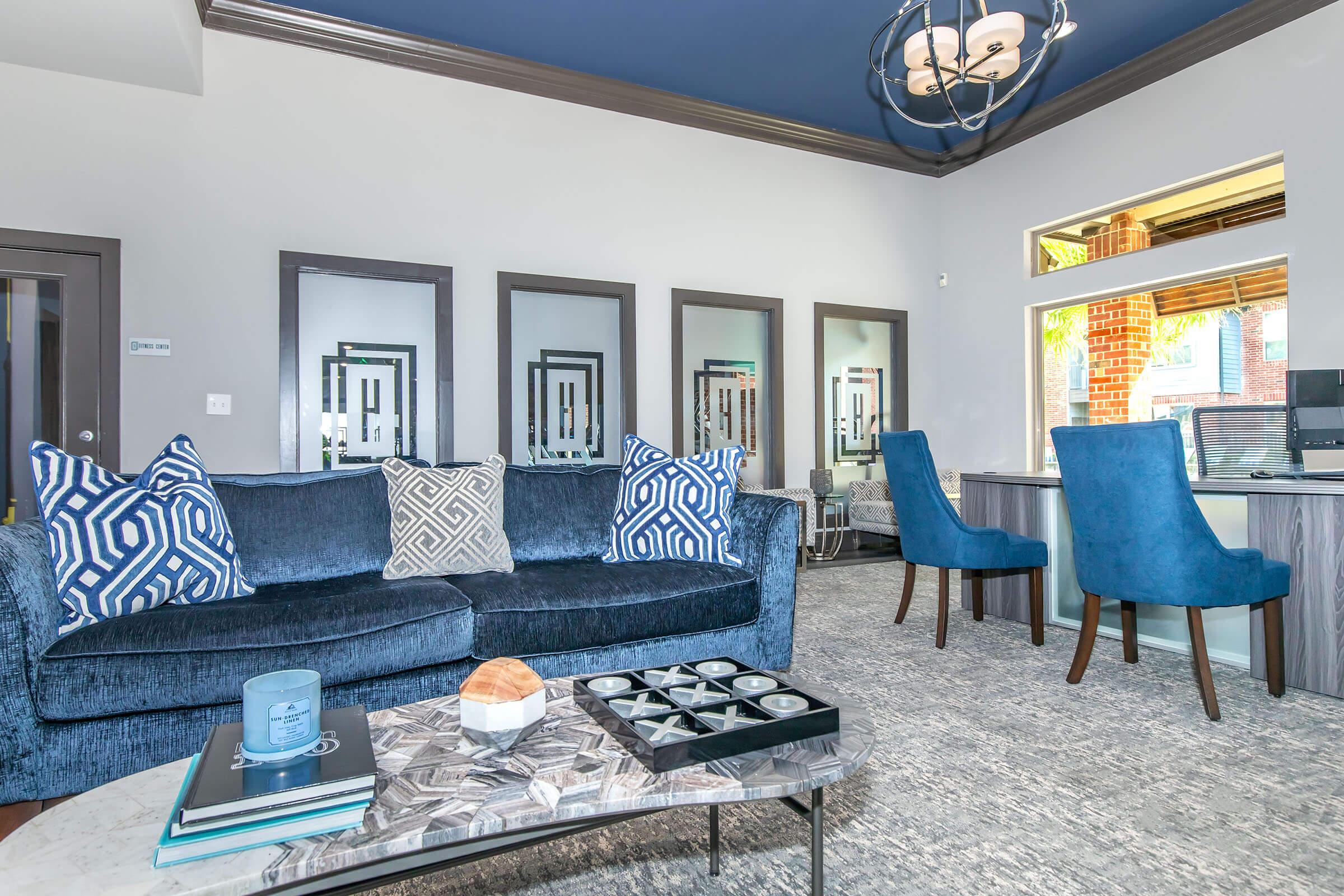 A modern living room featuring a blue velvet sofa with decorative pillows, a sleek coffee table with candles, and a patterned rug. The space includes a stylish dining area with blue chairs and large windows, complemented by contemporary artwork on the walls. The ceiling is painted dark blue, adding elegance to the decor.