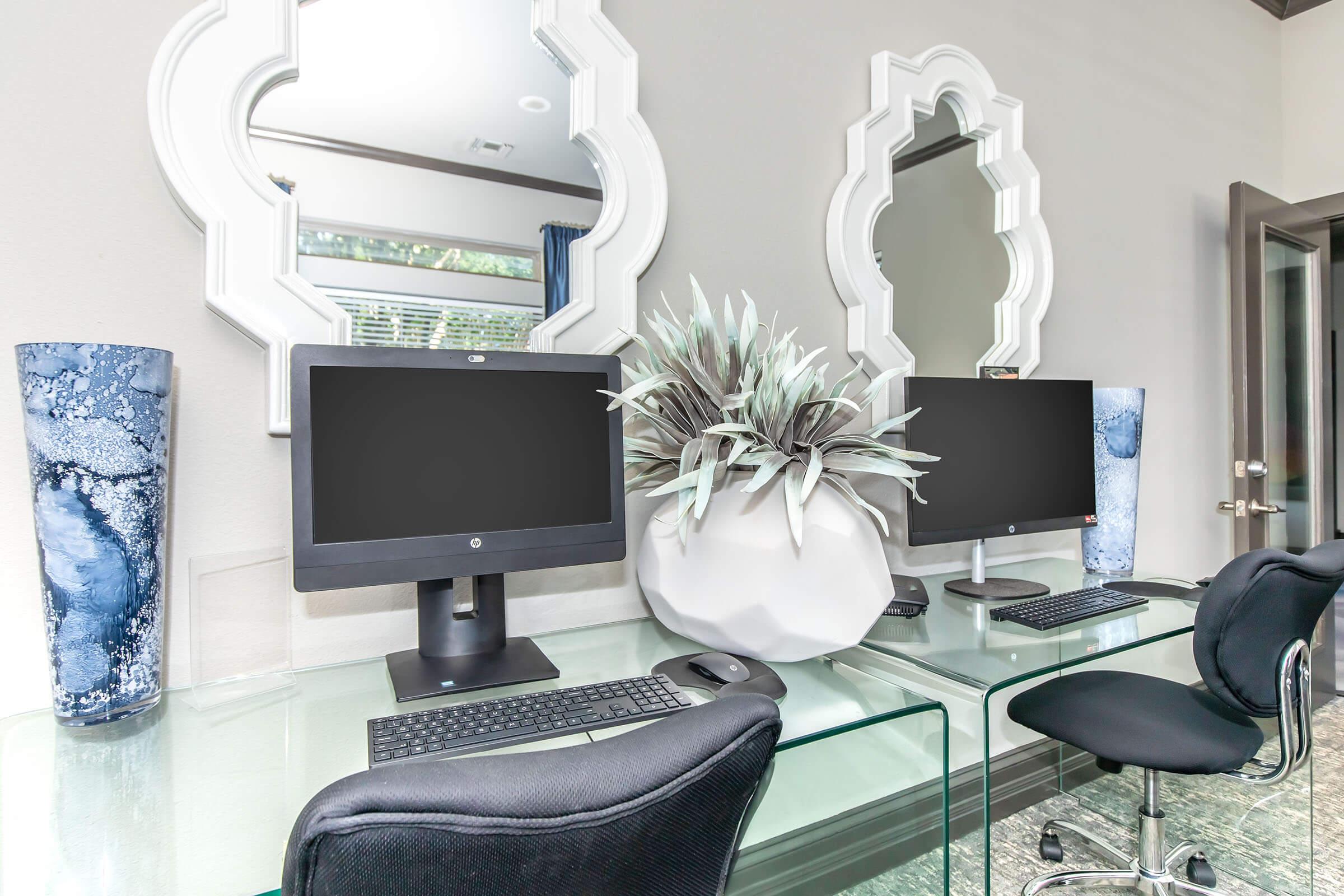 Two sleek computer workstations are set up on a glass desk, each featuring a black monitor and a keyboard. A large, modern white planter with greenery adds a touch of nature to the space. Behind the workstations, two ornate mirrors reflect the room's contemporary design, with blue decorative vases on either side.
