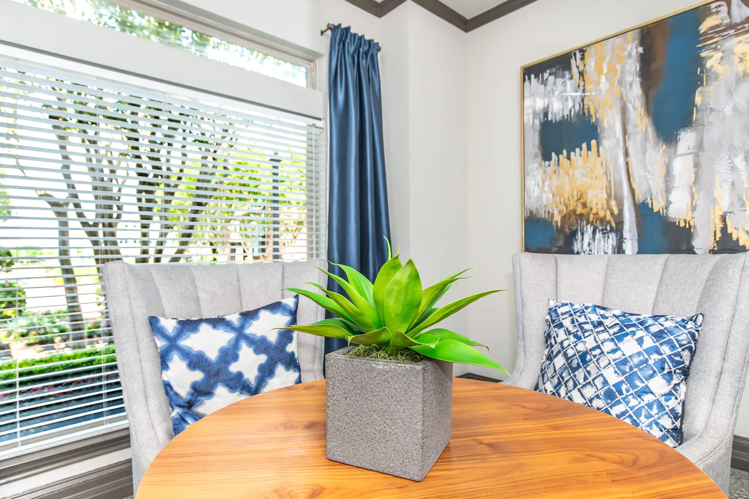 A cozy indoor space featuring a round wooden table with a potted green plant in a gray square pot. Two light gray armchairs with blue and white patterned pillows are positioned beside the table. Large windows with blinds provide natural light, and a modern artwork hangs on the wall, enhancing the ambiance.
