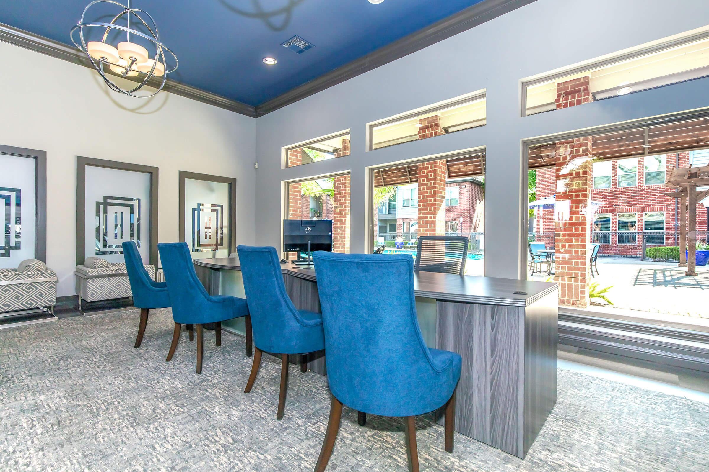 A bright and modern interior of a workspace featuring a long desk with several blue upholstered chairs. Large windows provide natural light and a view of an outdoor area. The ceiling is painted dark blue, and there are decorative light fixtures. The overall ambiance is inviting and professional.