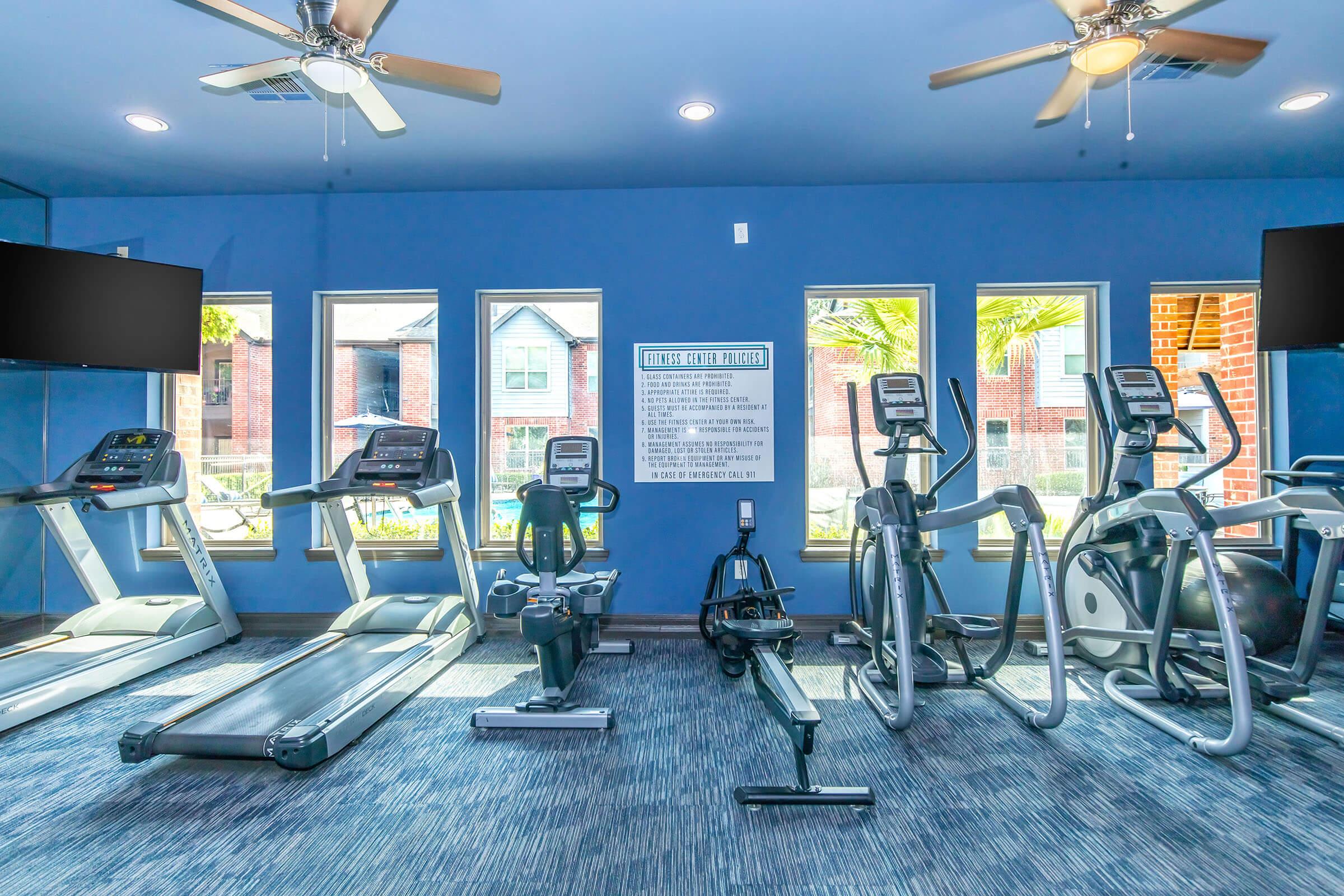 A modern fitness room featuring various exercise equipment, including treadmills, ellipticals, and a rowing machine. The room has large windows with natural light, ceiling fans, and two flat-screen TVs mounted on the walls. A motivational sign is displayed, and the flooring is a blue, textured carpet.