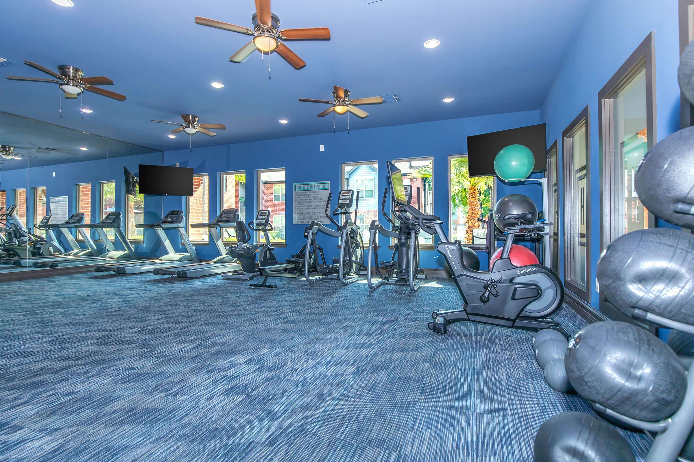 A modern fitness room featuring several exercise machines, including treadmills and stationary bikes, set against light blue walls and large windows. The space has a clean, airy feel with ceiling fans and colorful exercise balls in the corner, creating an inviting atmosphere for workouts.