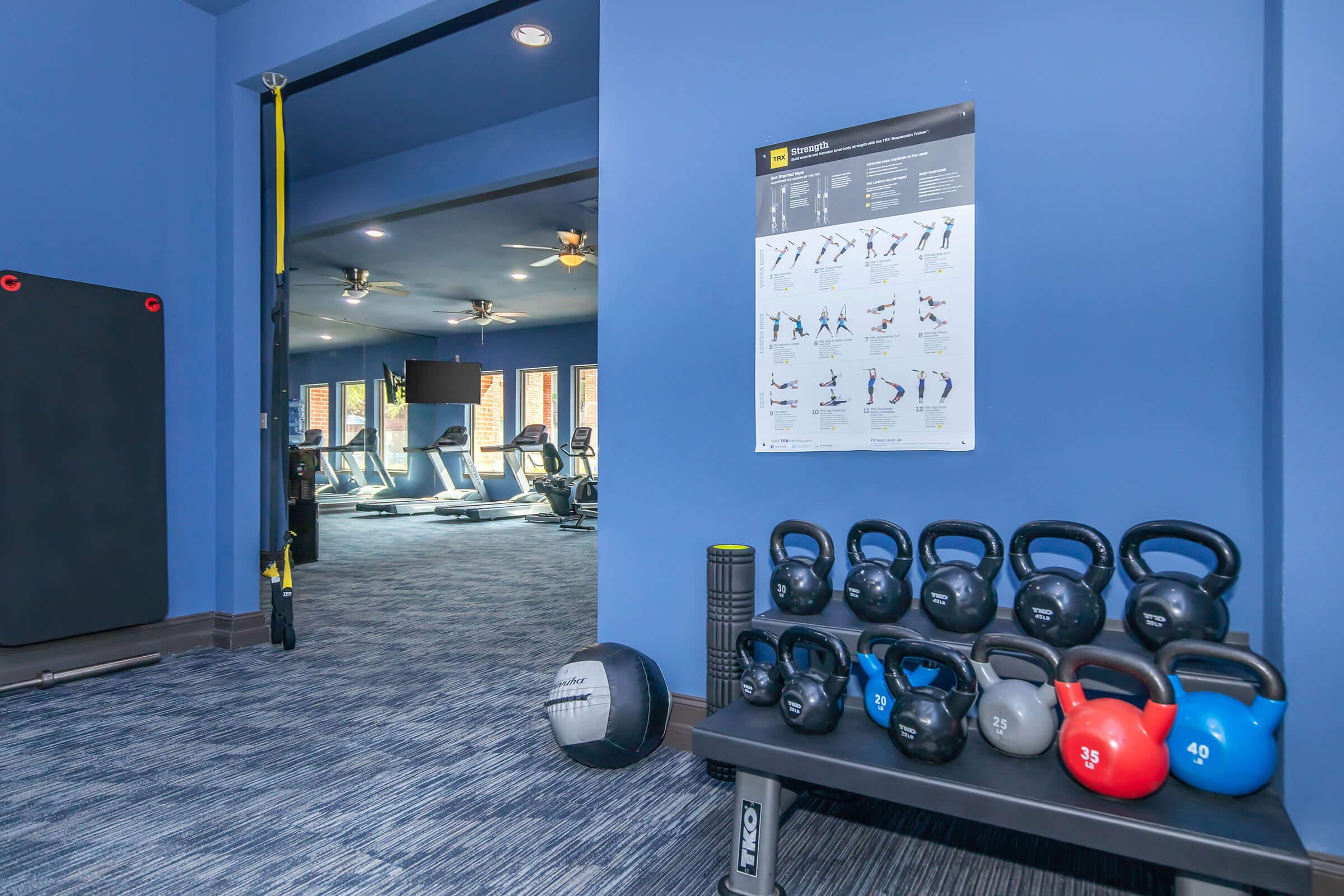 A well-equipped gym area featuring a rack of kettlebells of various weights, a medicine ball, and a foam roller on a black bench. A poster on the wall displays exercise instructions. The background shows cardio equipment in a spacious, modern gym setting with blue walls and good lighting.