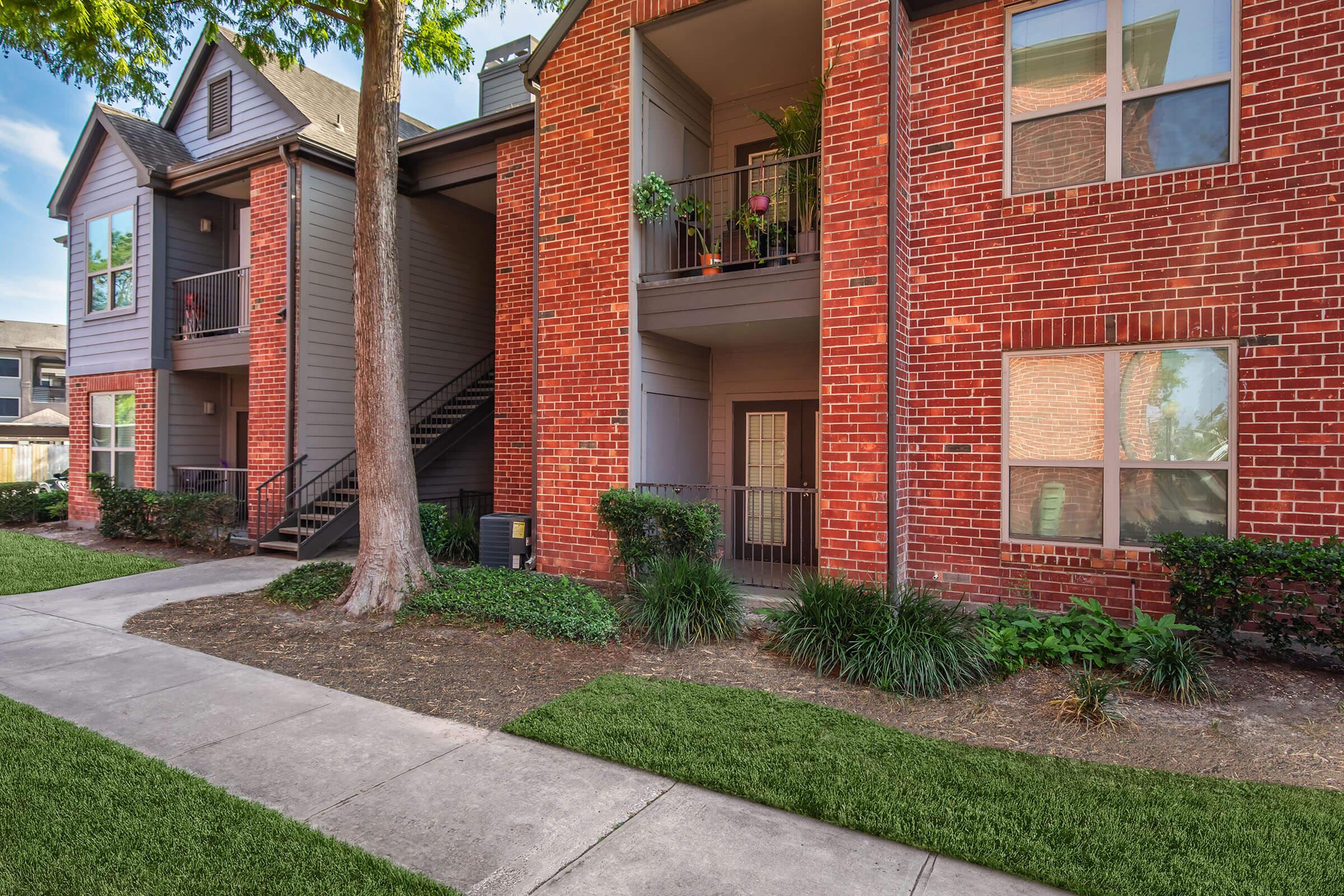 A view of a two-story apartment building made of red brick and gray siding. The building features balconies with potted plants, surrounded by greenery and trees. A concrete pathway leads to the entrance, with well-maintained landscaping lining the walkway.