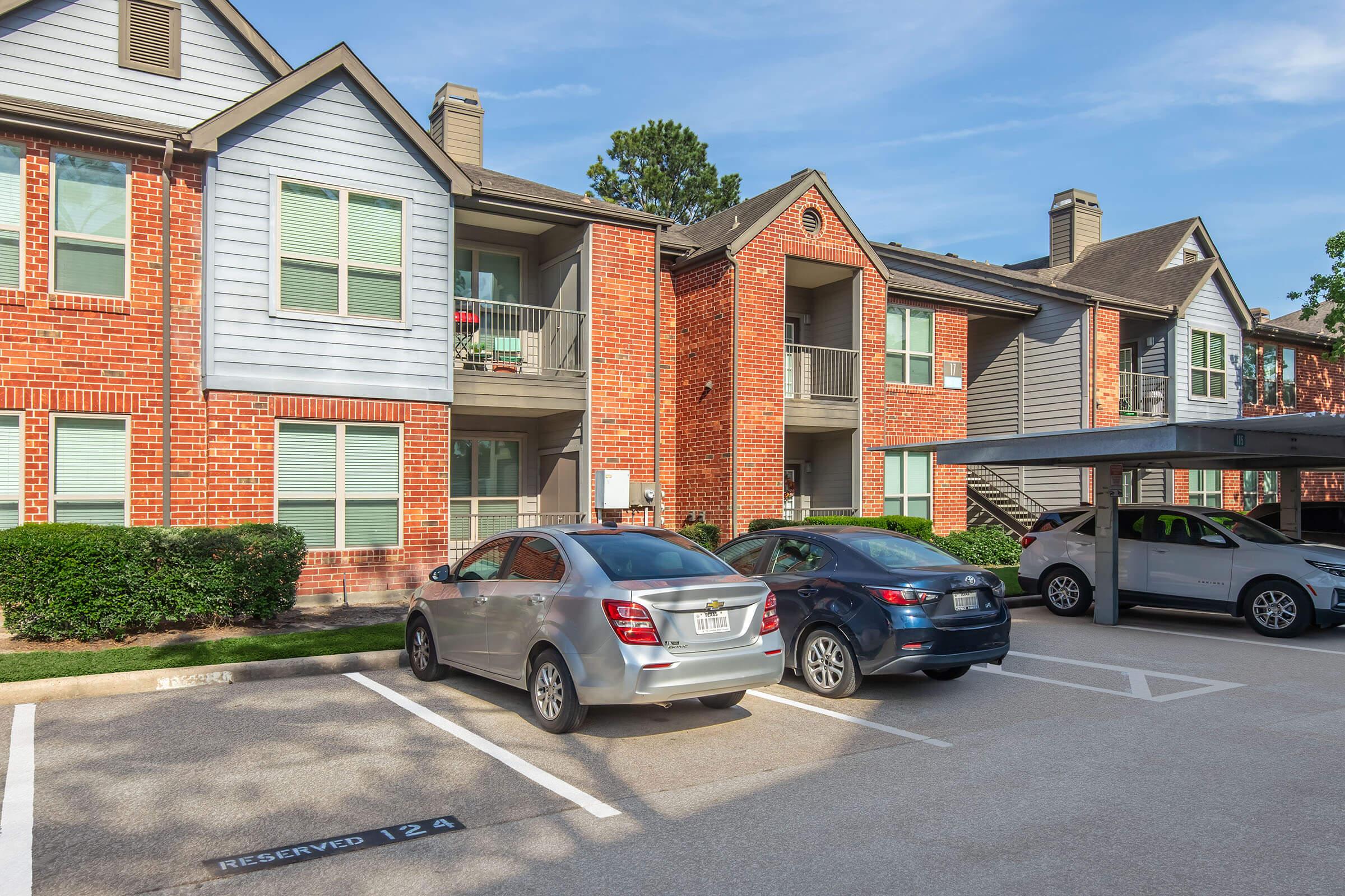 View of an apartment complex featuring two buildings with brick and wood siding. Several parked cars are in the foreground, with designated parking spots visible. The scene is set against a clear blue sky, with greenery surrounding the buildings, suggesting a well-maintained residential area.