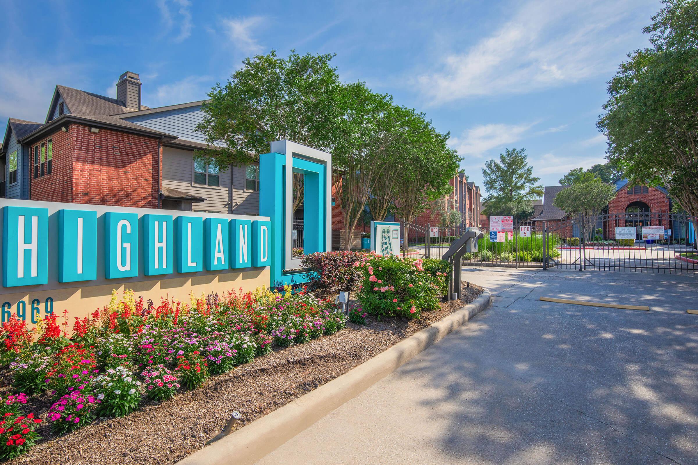 A colorful entrance sign for "Highland," surrounded by well-maintained landscaping featuring blooming flowers and neatly trimmed shrubs. The entrance leads to a gated community with buildings visible in the background under a clear blue sky.