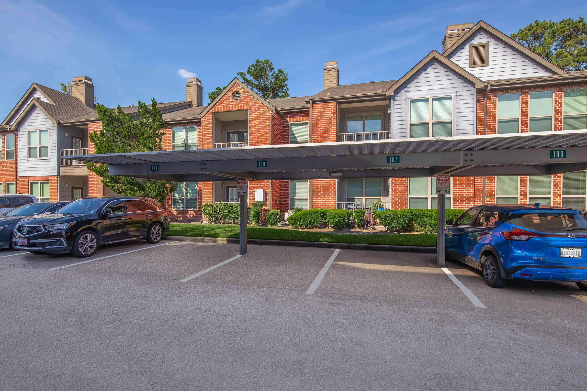 A residential apartment complex featuring several units with brick and siding exteriors. In the foreground, there is a covered parking area with parked cars, including a black car and a blue car. Lush green landscaping surrounds the building under a clear blue sky.