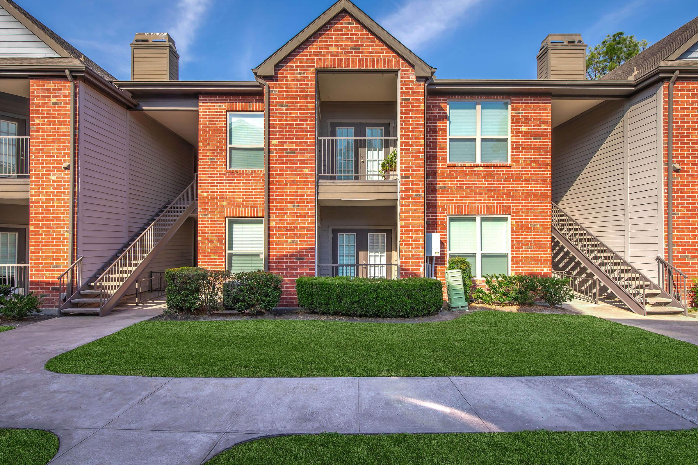 A two-story apartment building with a brick facade. The entrance features a covered porch with access to the upper and lower floors. Lush green grass surrounds the walkway, and there are stairs leading to the upper level on both sides. The sky is clear with a few clouds.