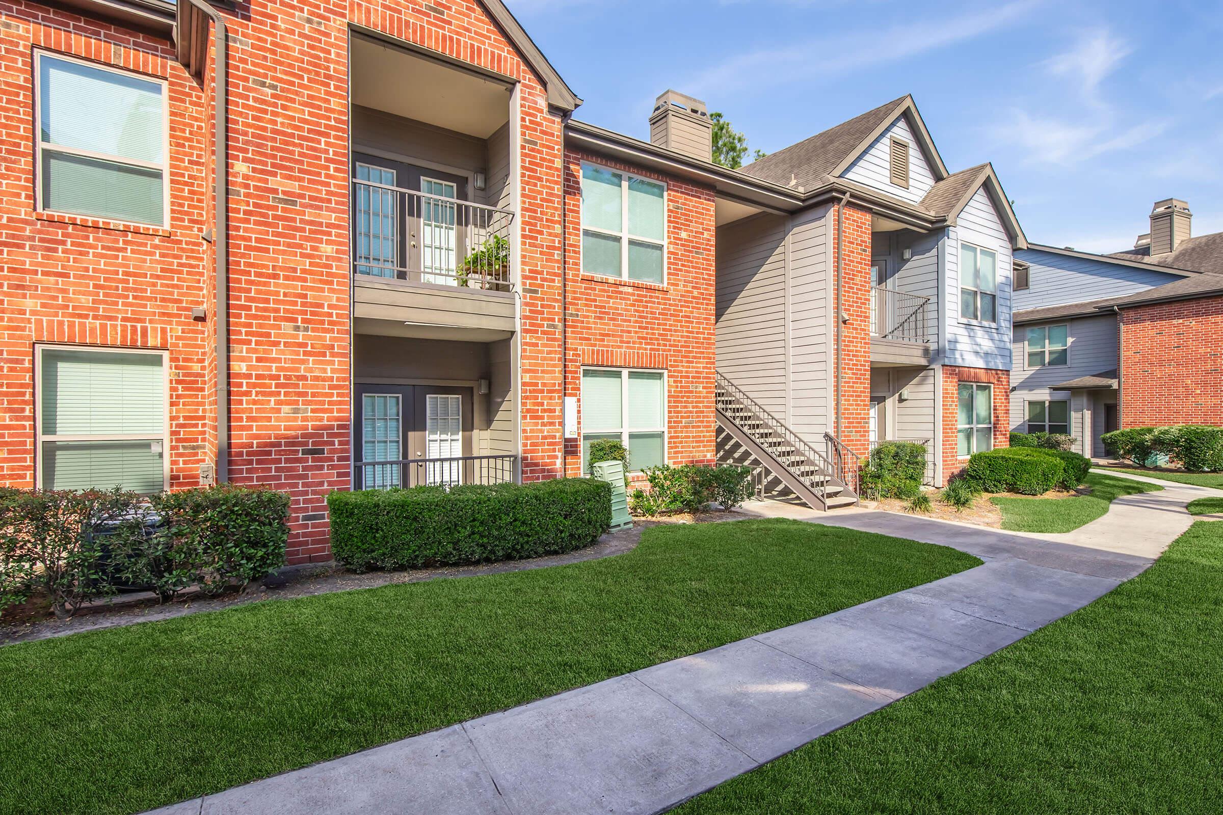 A well-maintained apartment complex featuring red brick and gray siding buildings. The landscape includes neatly trimmed bushes and grass, with a walkway leading past the units. Balconies are visible on the upper levels, and the setting is bright with clear blue sky.