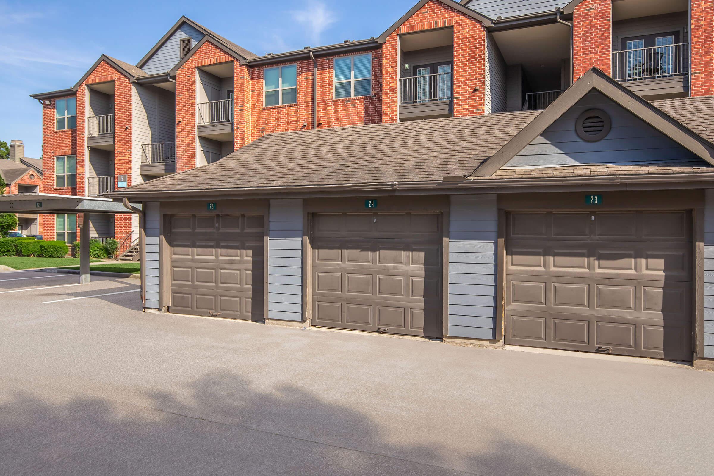 A residential building featuring multiple units with brick and gray siding. In the foreground, three garage doors are lined up, each with a numbered label. The setting includes a paved parking area, and the sky is clear with visible sunlight.