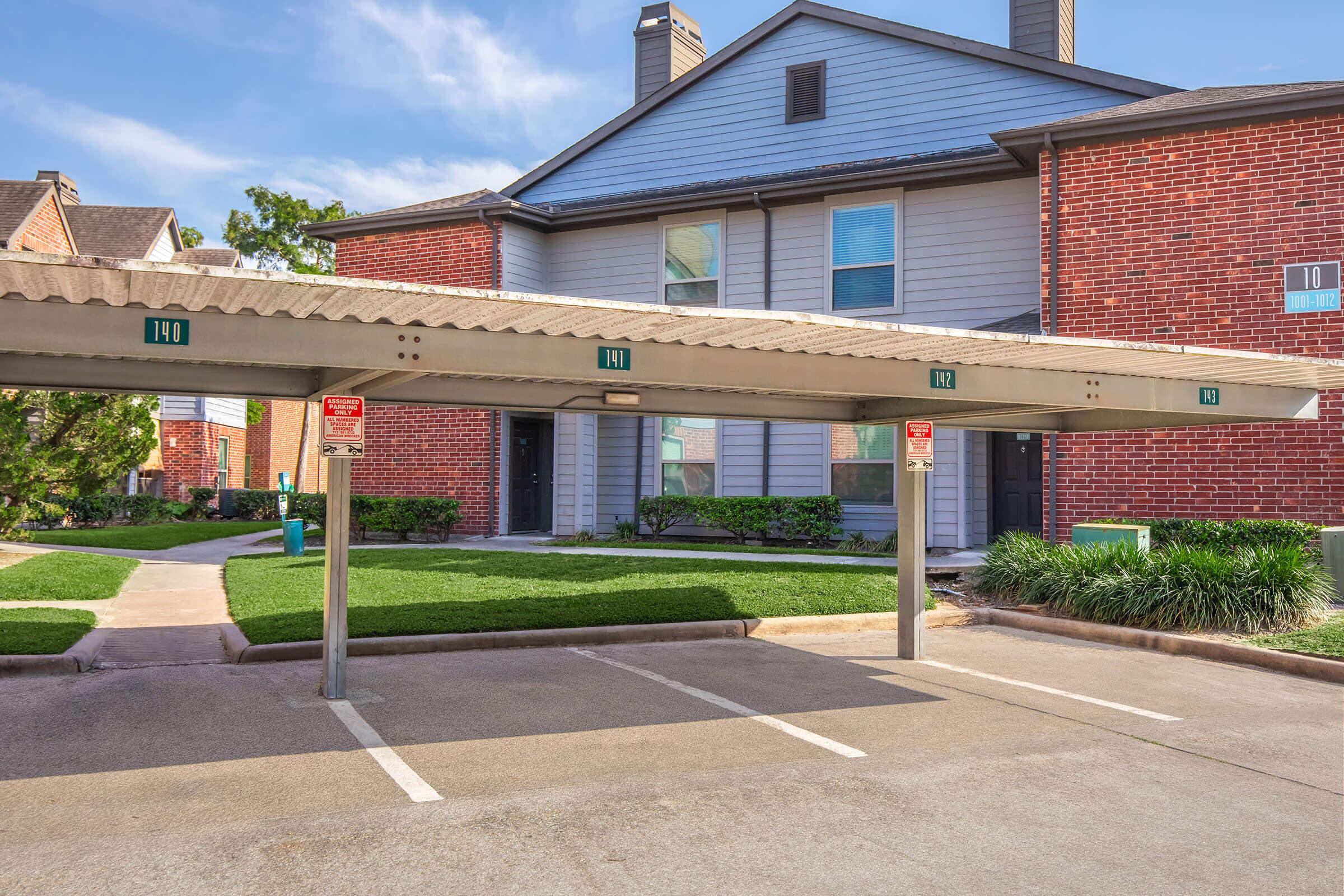 A covered parking area with two empty spots in front of a red brick apartment building with gray siding. Green grass and shrubs are visible along the walkway leading to the entrance. The sky is clear with a few clouds. Signage indicates reserved parking.