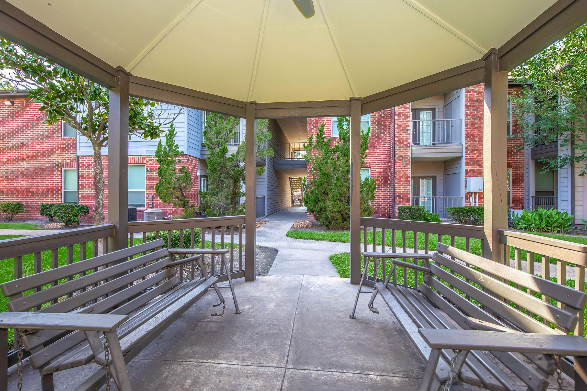A peaceful outdoor gazebo with wooden benches, surrounded by lush greenery and brick buildings. A walkway runs through the center, leading to more buildings in the background, creating a tranquil atmosphere for relaxation and socializing.