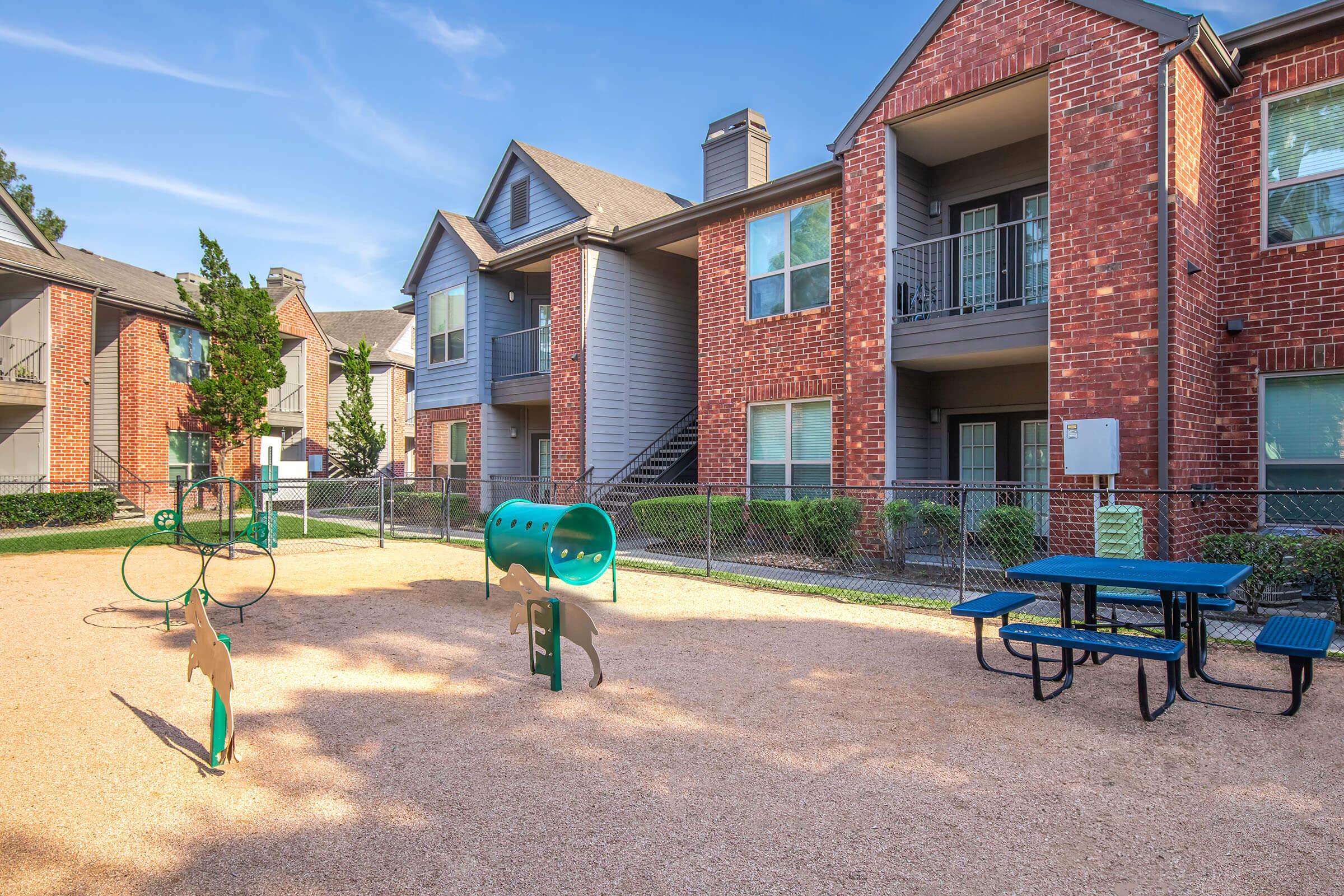 View of a fenced outdoor playground area featuring play equipment, a picnic table, and brick apartment buildings in the background. The ground is covered in gravel, and there are trees and greenery surrounding the area under a clear blue sky.