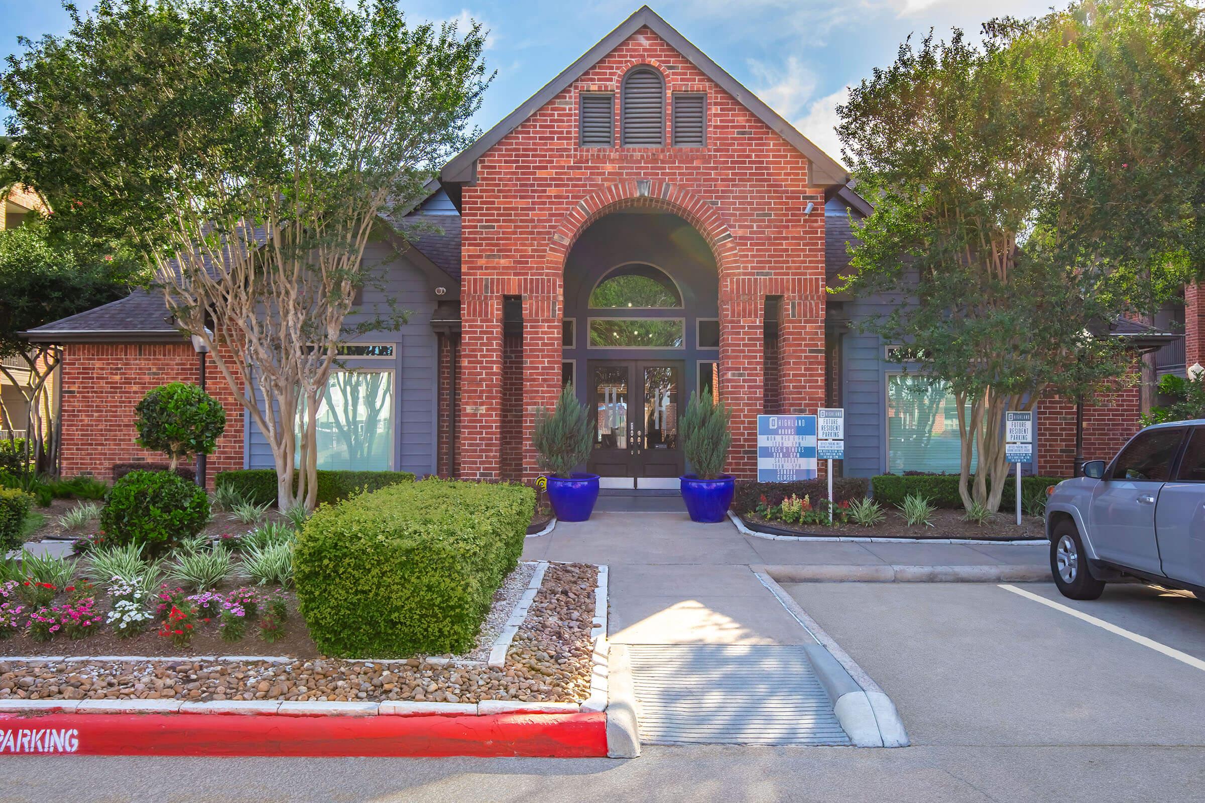 A brick building with large windows framed by shrubs and trees. The entrance features a prominent arch and is flanked by blue planters. A landscaped area with gravel and plants is visible in front, alongside a parking area with a car parked. The scene is bright, suggesting a welcoming atmosphere.