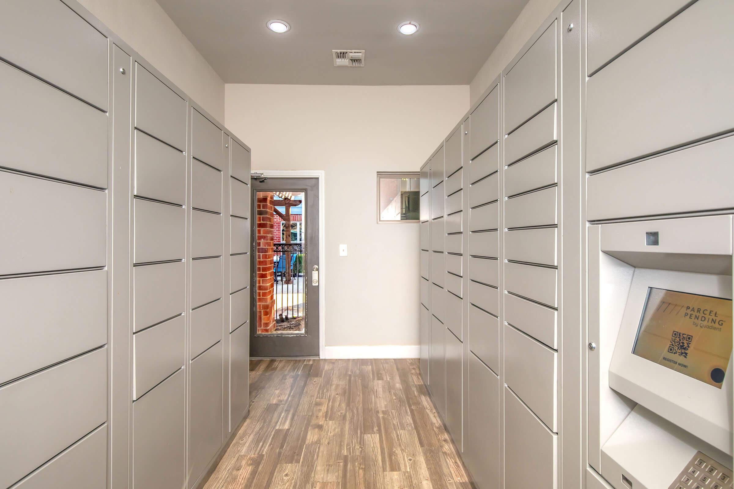 A hallway lined with metallic gray parcel lockers on either side, leading to a glass door that opens to an outdoor area. A digital screen for parcel handling is visible on the right side, with wooden flooring beneath bright overhead lights.